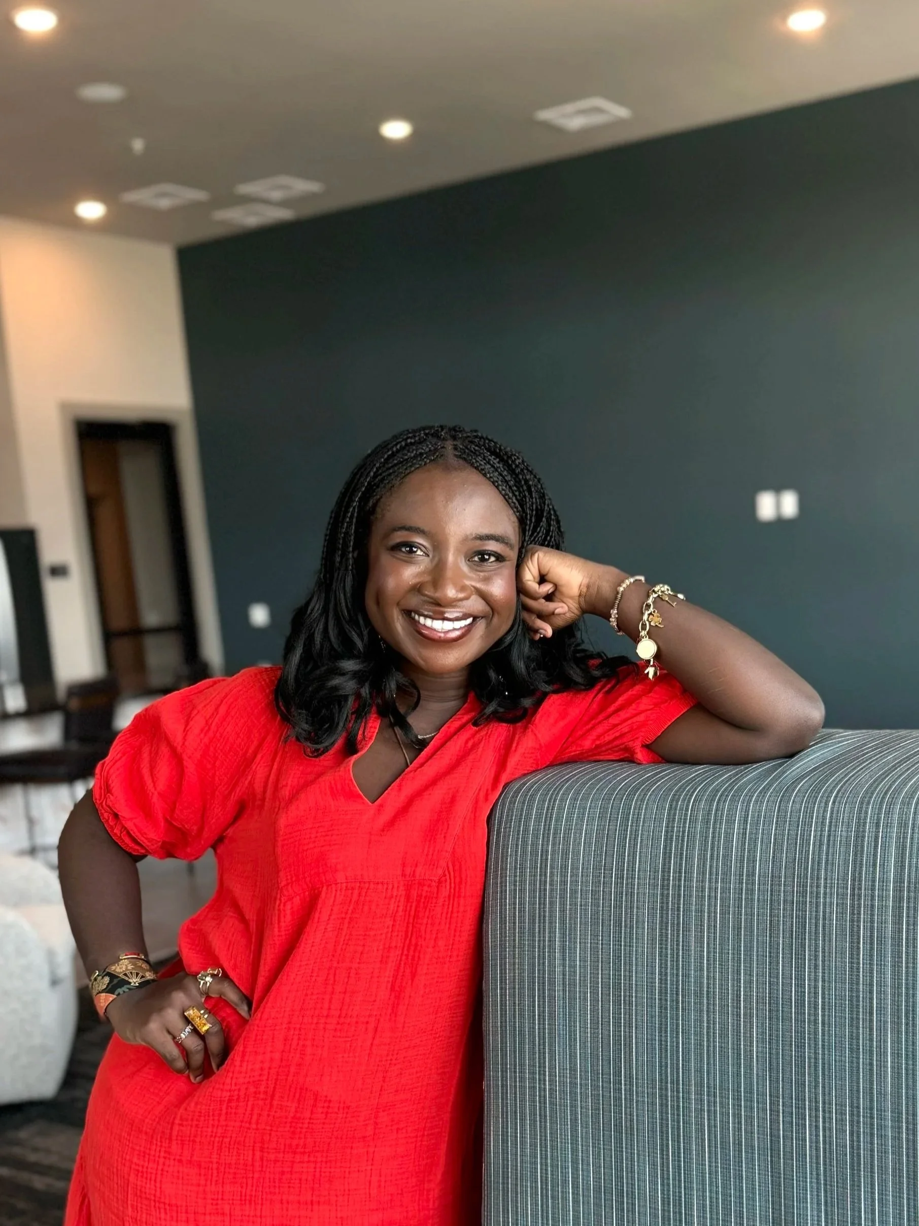 A smiling Black woman with braided hair wearing a red dress, sitting indoors with her elbow resting on a piece of furniture.