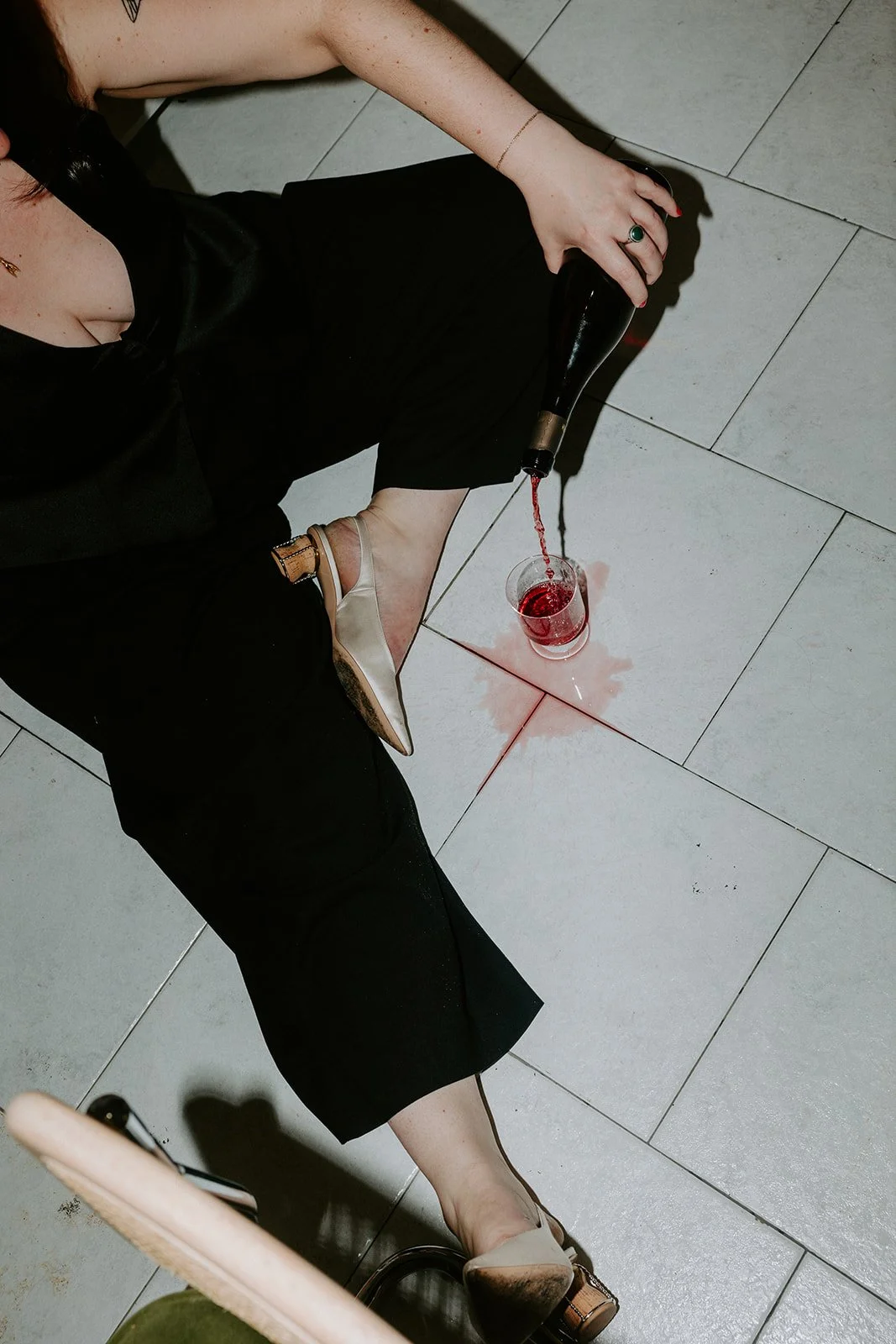 A woman sitting on the floor pouring red wine from a bottle into a glass, with spilled wine on the tile floor near her.