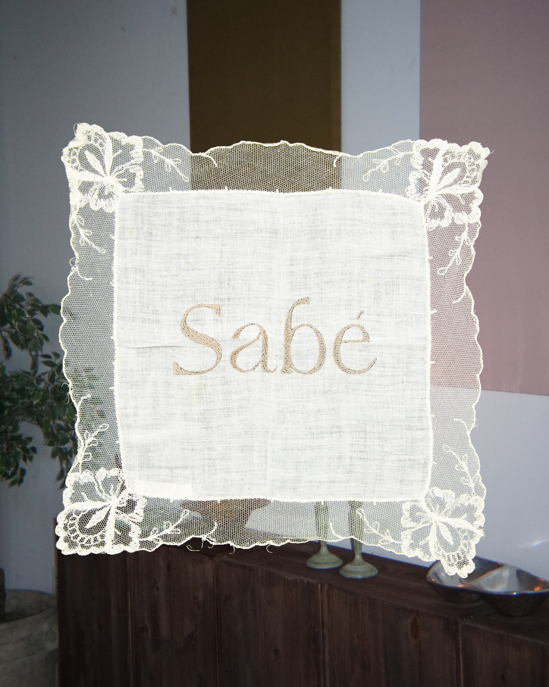 A lace-trimmed white fabric sign with the word "sabe" embroidered in the center, hanging indoors with a wooden cabinet and potted plant in the background.