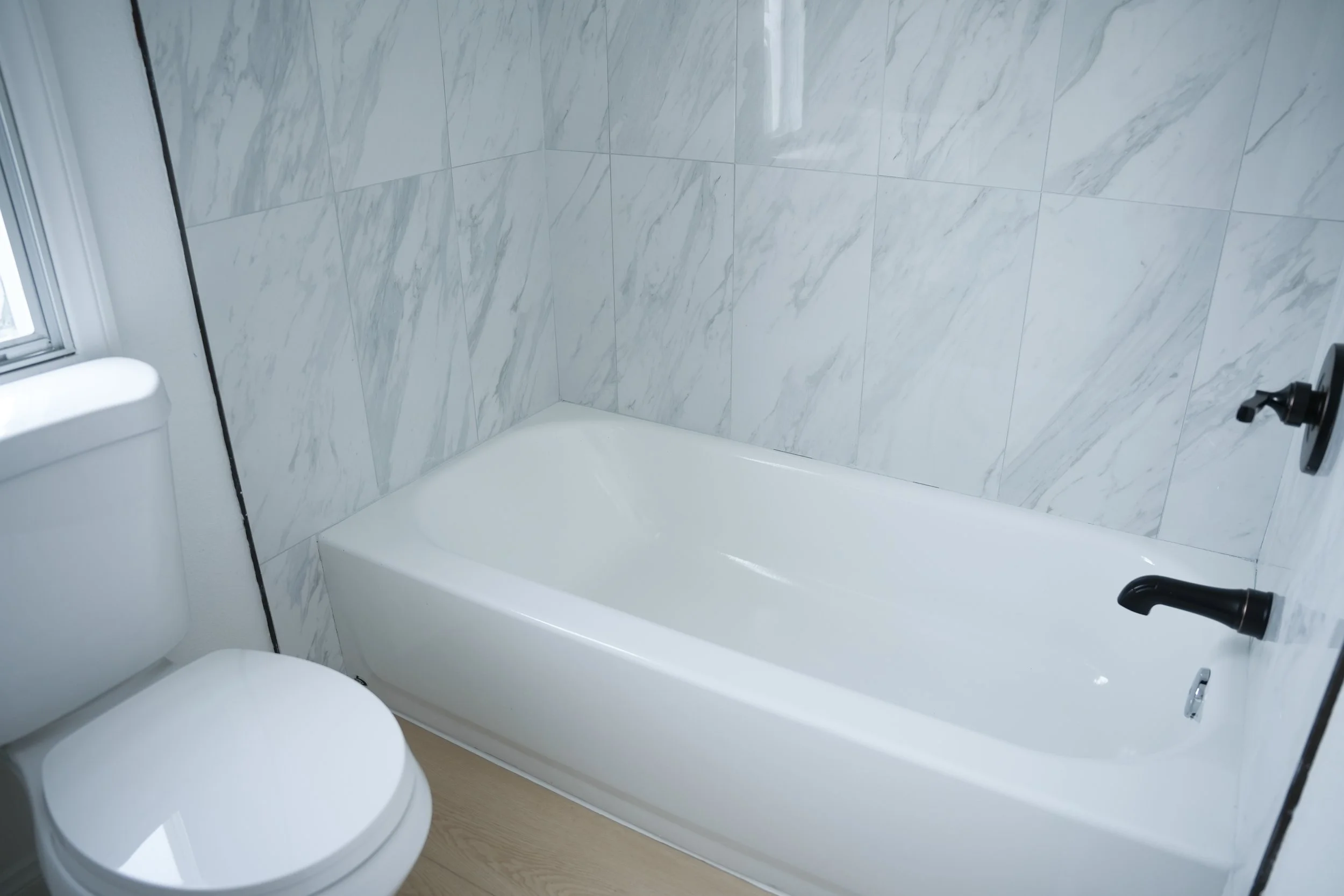 Bathroom with white bathtub and toilet, marble tile walls, and black fixtures.