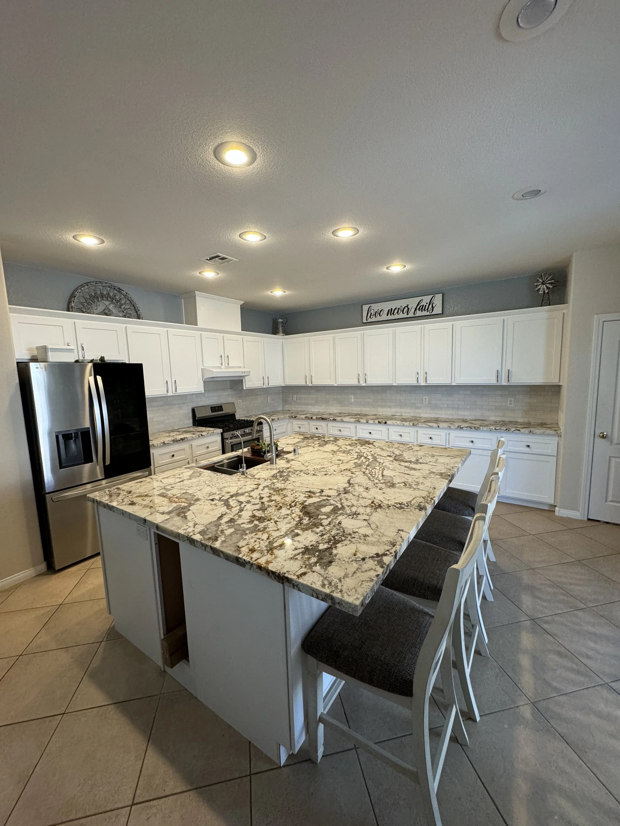 Modern kitchen with a large island and barstools, granite countertops, stainless steel refrigerator, and white cabinets.