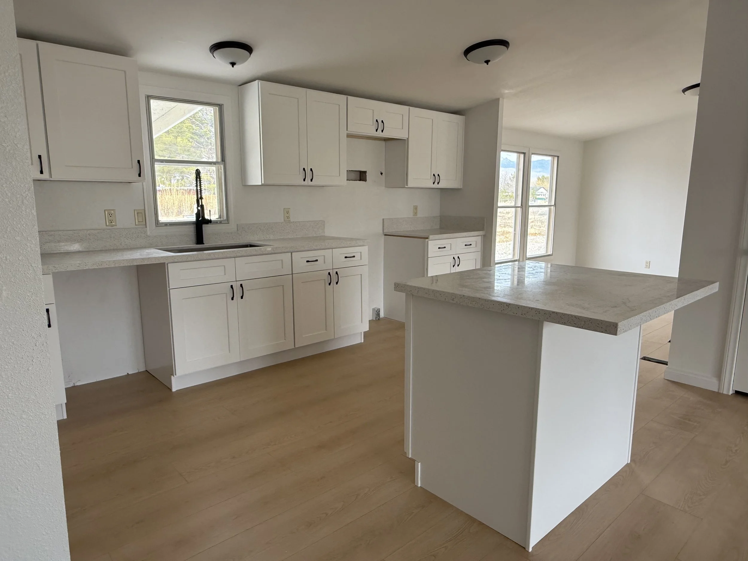 Modern kitchen interior with white cabinets, light wood flooring, and a central island. Features a window, black faucet, and minimalist design.