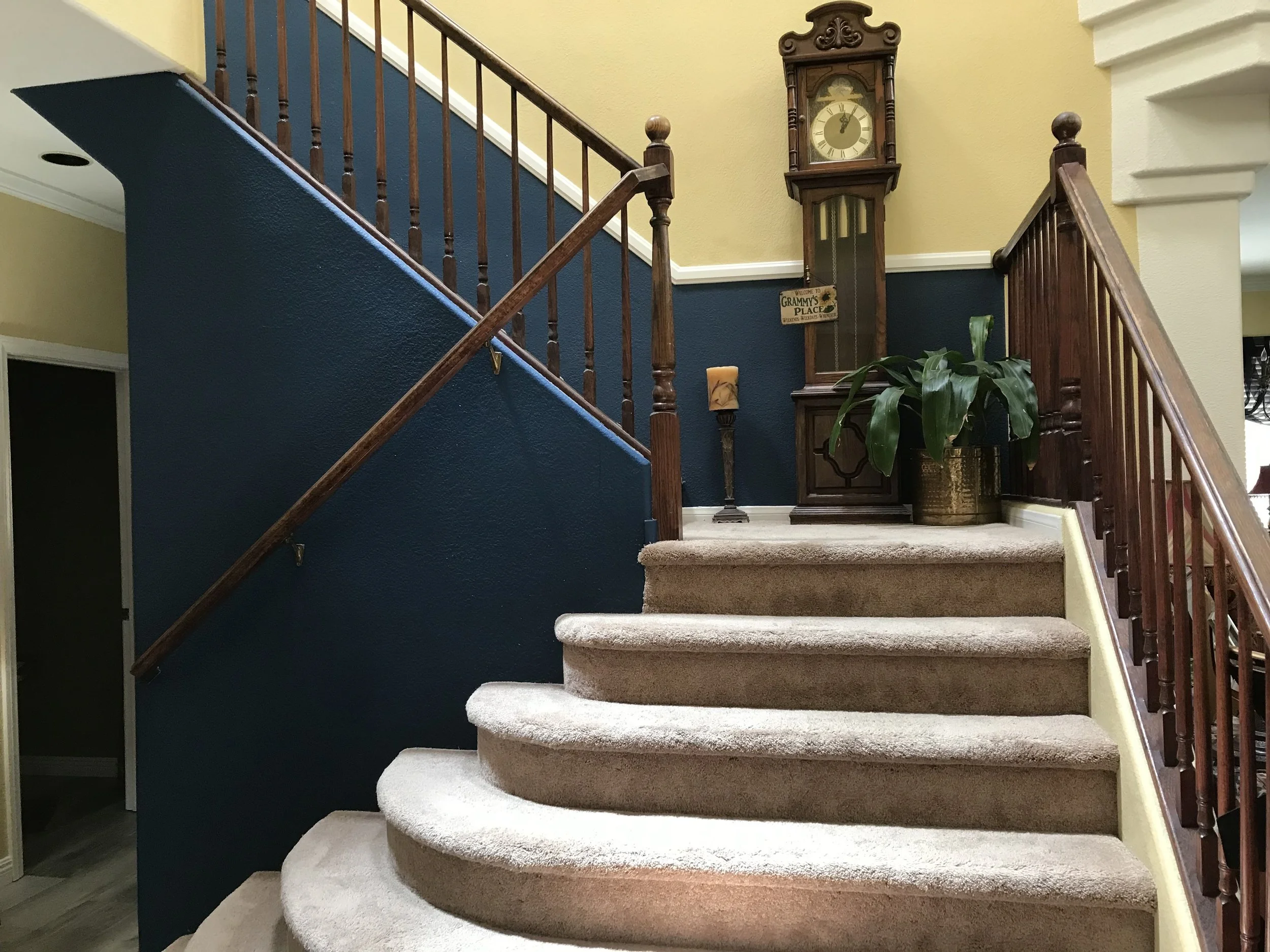 Interior staircase with beige carpet, dark blue and yellow walls, wooden railings, a tall antique clock, a plant in a brass pot, and a candle on a stand.