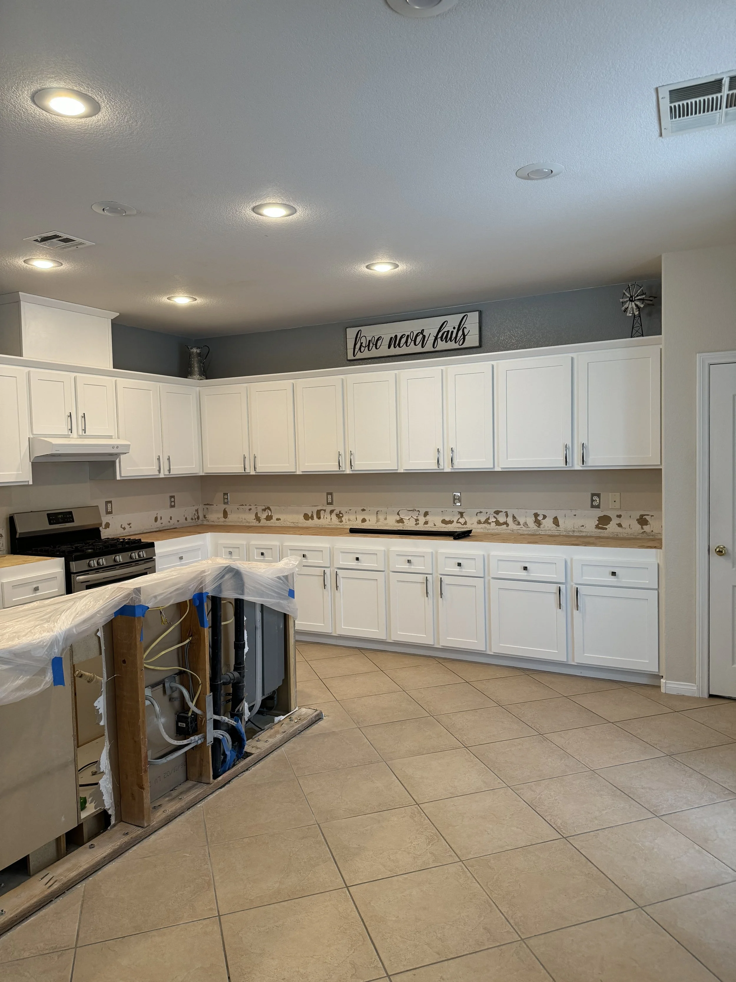 Kitchen under renovation with white cabinets, tiled floor, and exposed plumbing. "Love never fails" sign on top of cabinets.