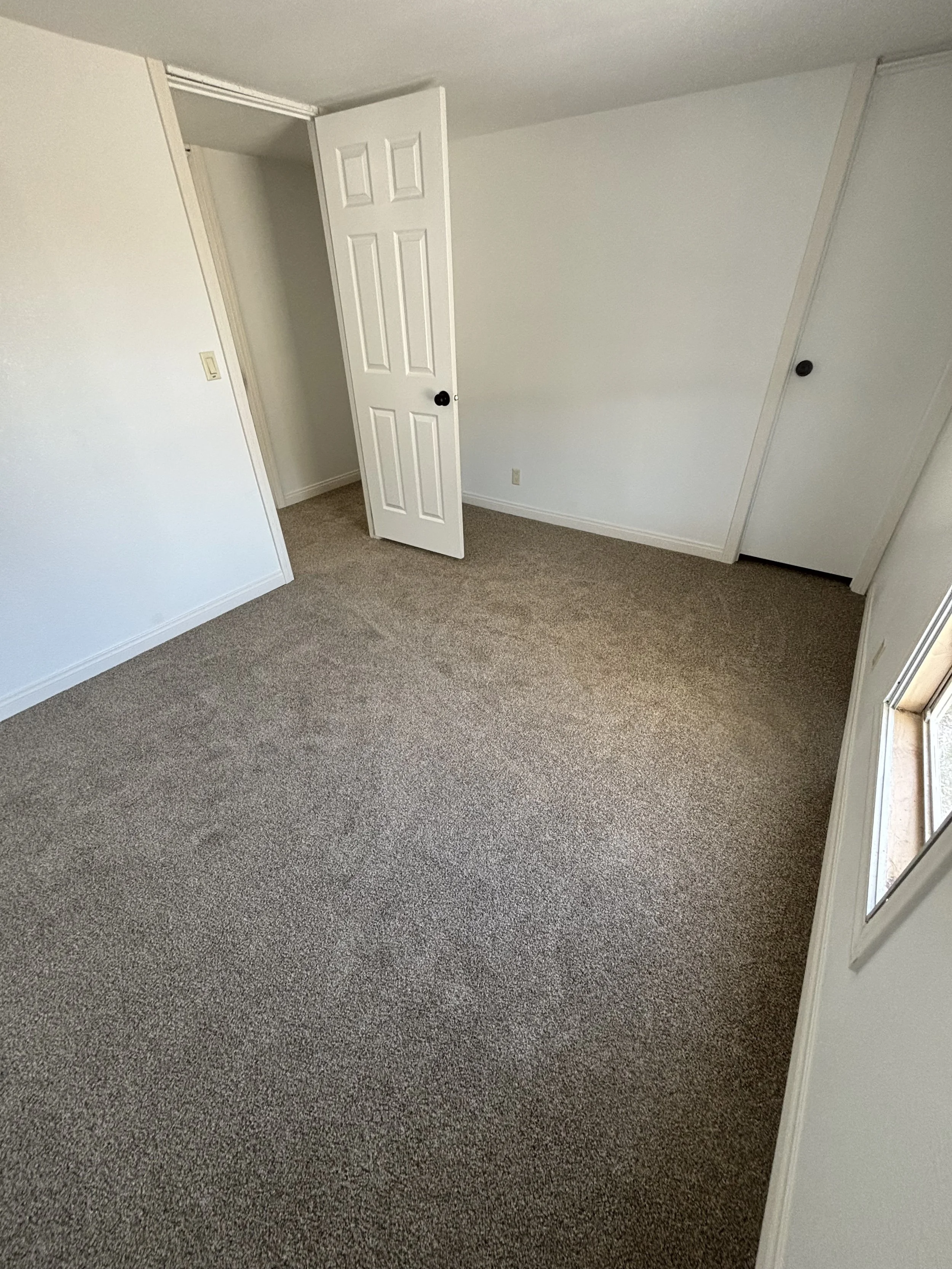 Empty white room with beige carpet, open door, and small window.