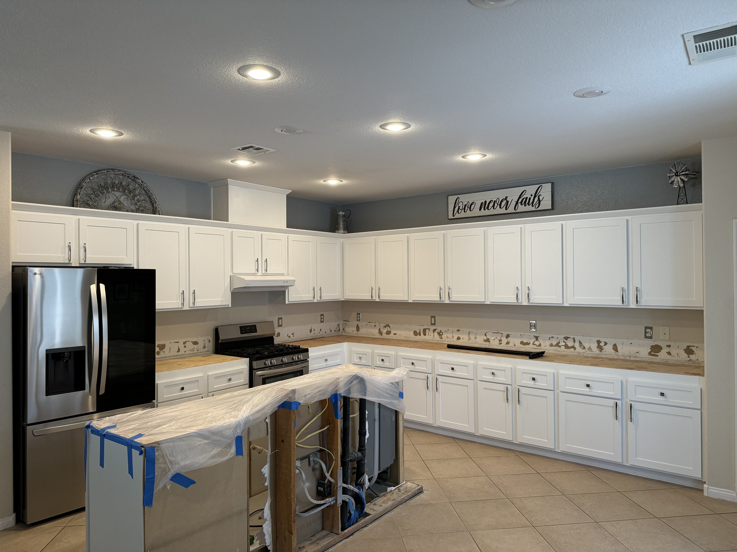 Modern kitchen under renovation with white cabinets, stainless steel refrigerator, and a partially installed kitchen island. Overhead lights illuminate the space, which includes a "Love never fails" sign on the wall.