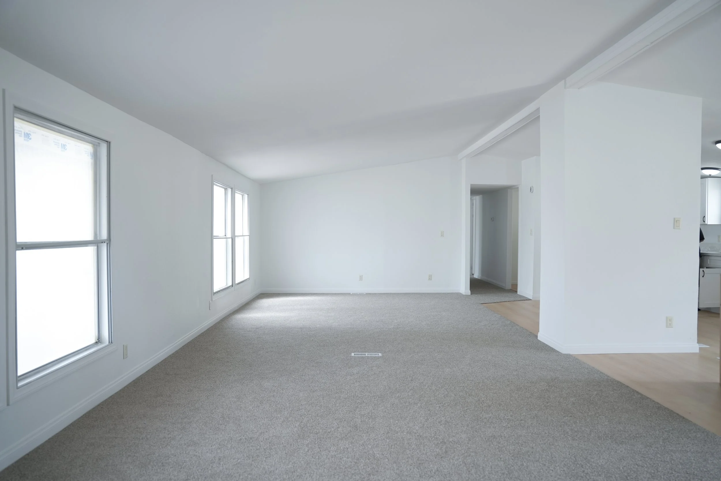 Empty living room with carpeted floor and large windows, leading to a hallway and a kitchen area with wooden flooring.