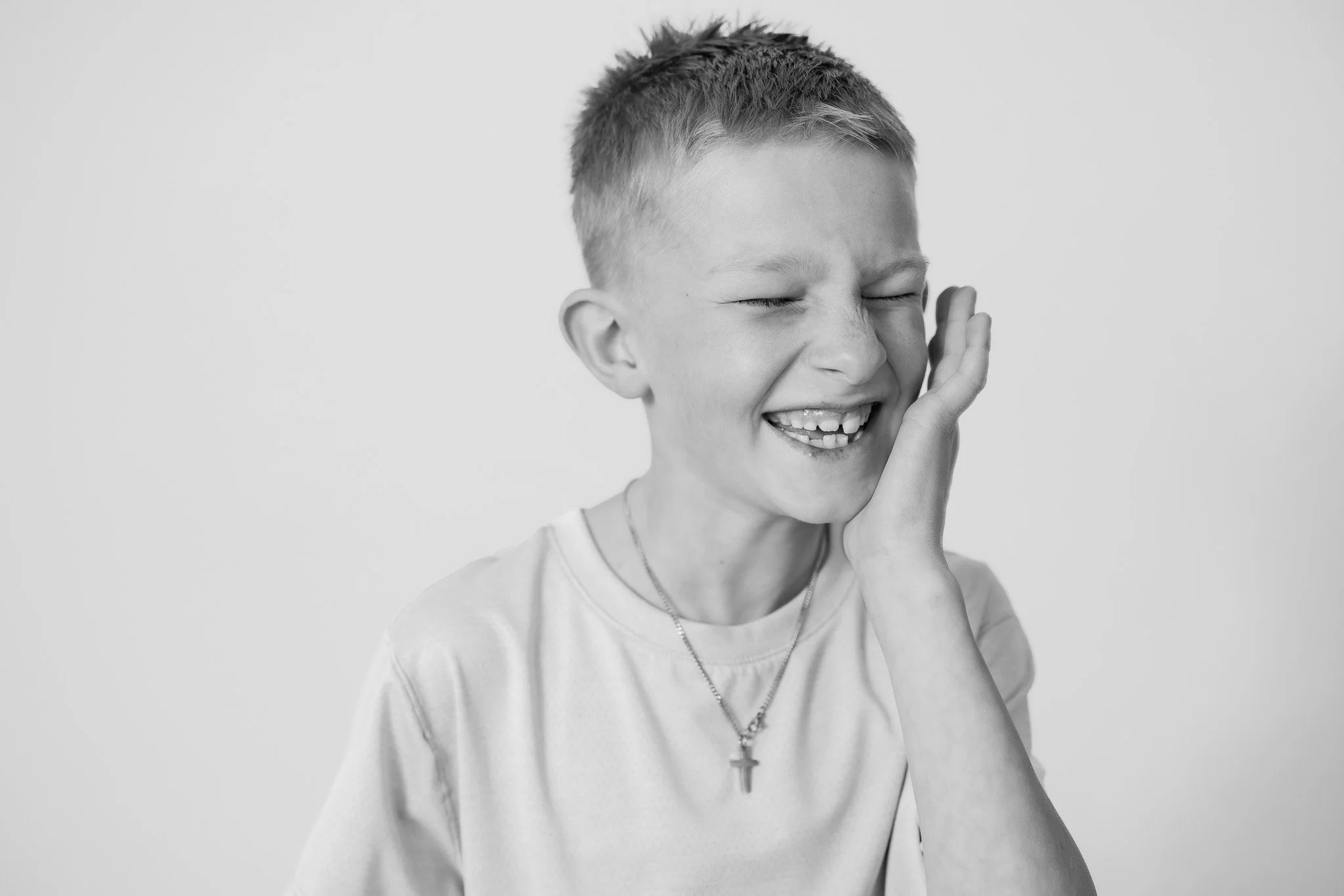 A young boy with short hair is smiling and closing his eyes, touching his cheek with his hand. He is wearing a light-colored t-shirt and a necklace with a cross pendant, and appears to be happy.