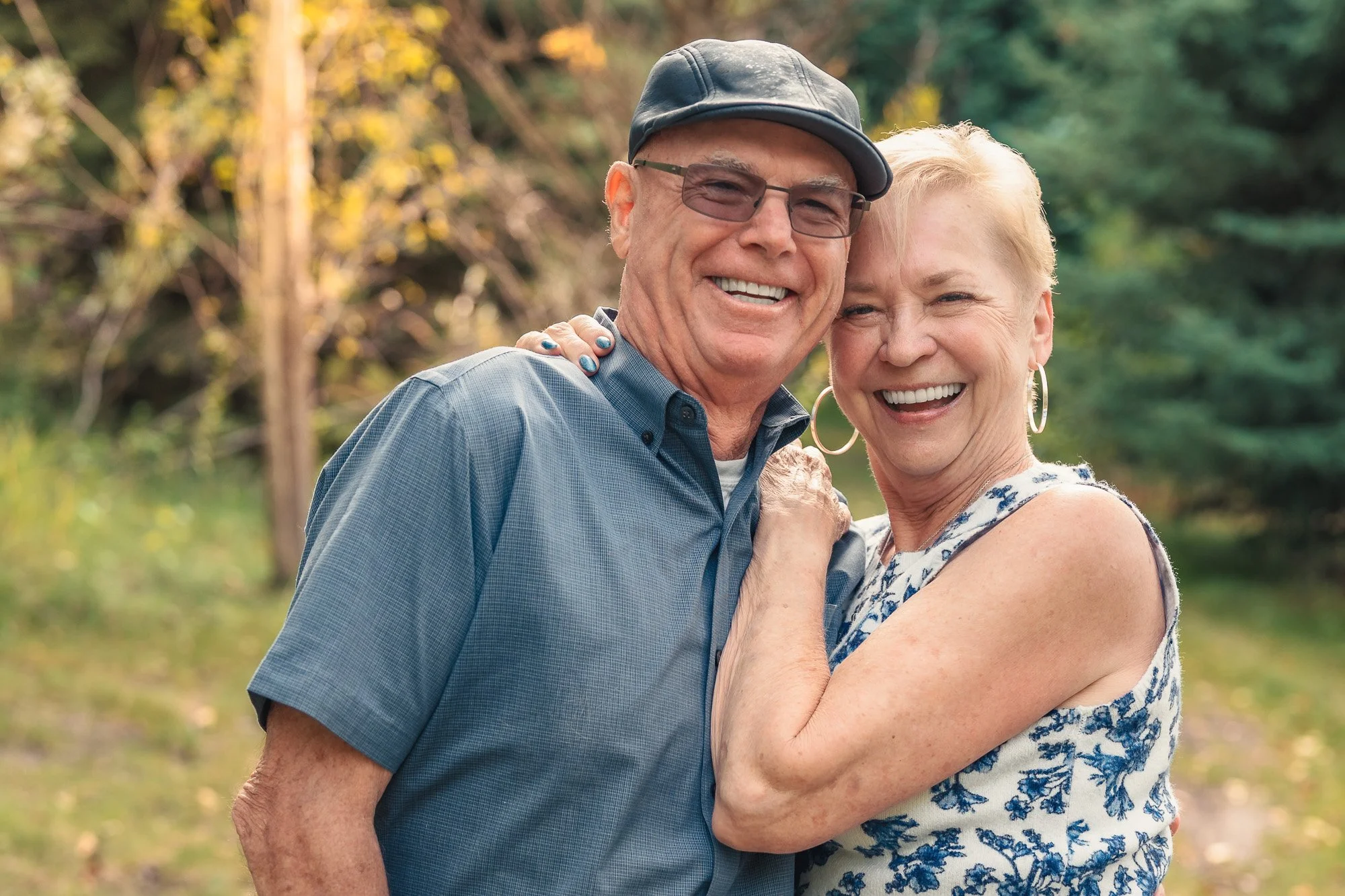 An elderly couple smiling and hugging outdoors in a park with green trees and autumn-colored foliage.
