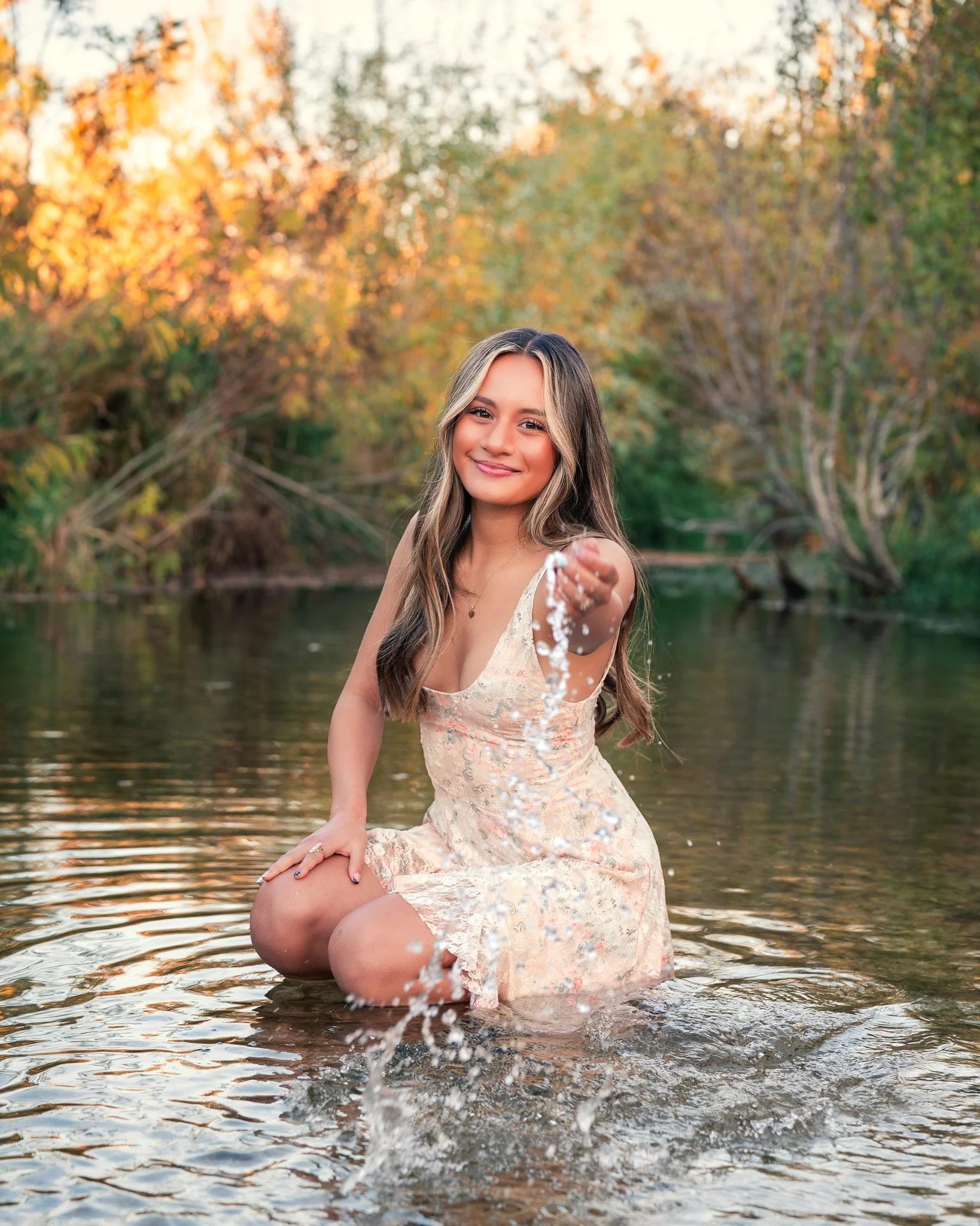 High school senior girl in floral dress kneeling in shallow river throwing water at sunset during fall senior pictures session with autumn foliage.