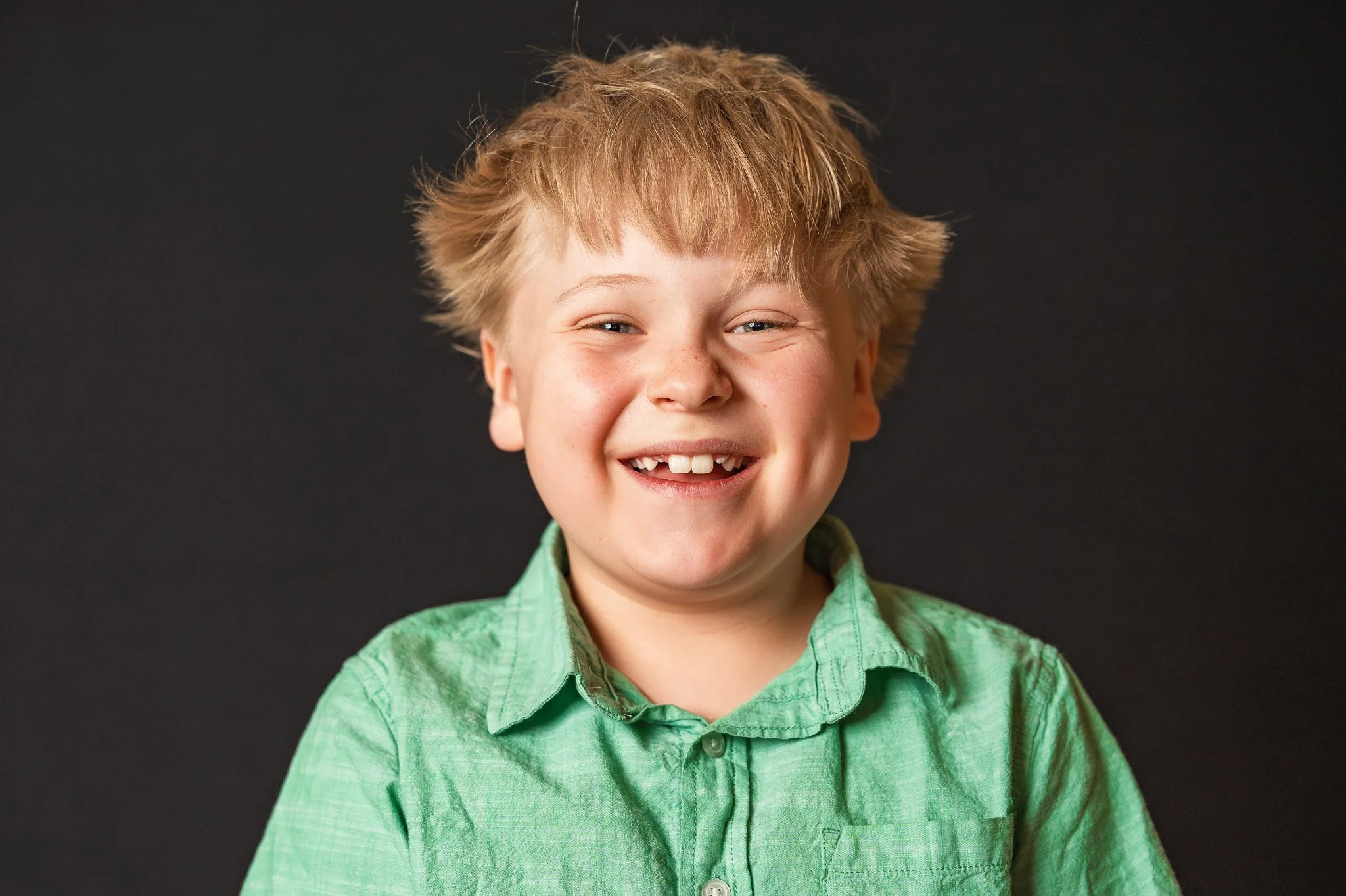 A boy with light skin, messy blonde hair, and a big smile showing missing front teeth, wearing a light green shirt against a dark background.