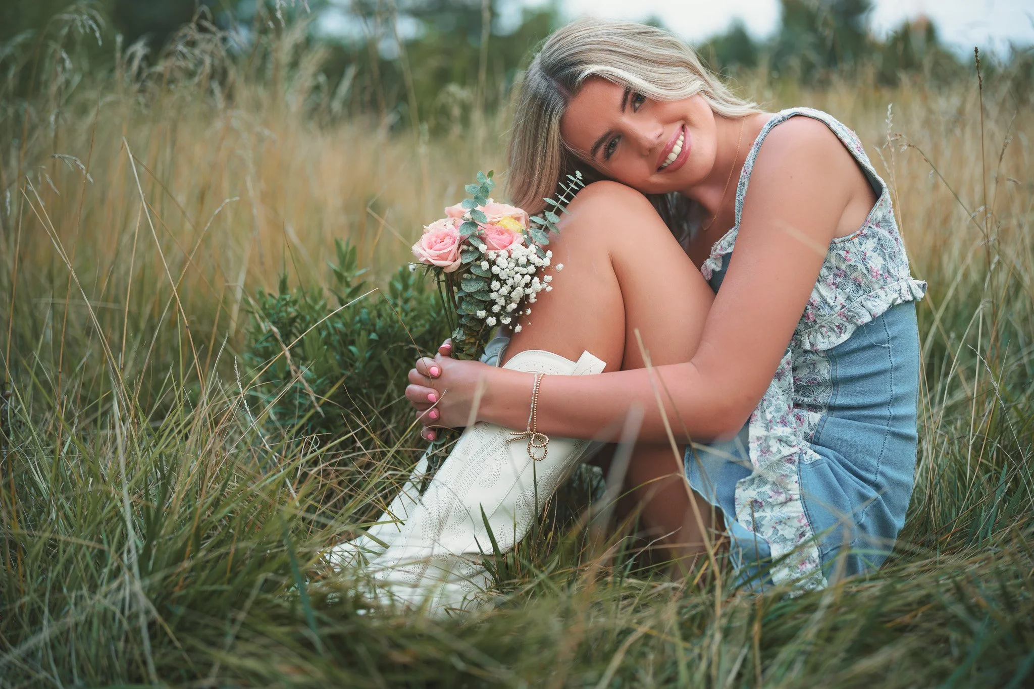Smiling high school senior girl sitting in tall grass holding pink roses and baby's breath bouquet at Kathryn Albertson Park during Boise senior pictures session.