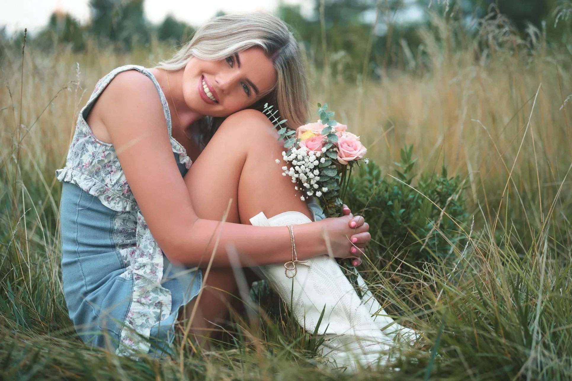 A woman with blonde hair smiling while sitting on the grass and holding a bouquet of pink and white flowers, with a man behind her hugging her and resting her head on his shoulder in a grassy field.
