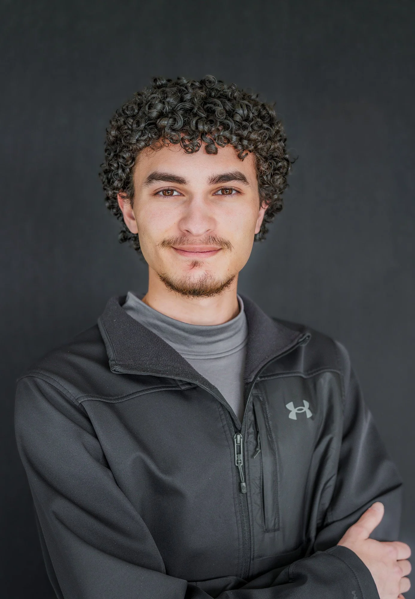 Close-up of a young man with curly dark hair, brown eyes, light facial hair, wearing a black Under Armour jacket and gray shirt against a dark background.
