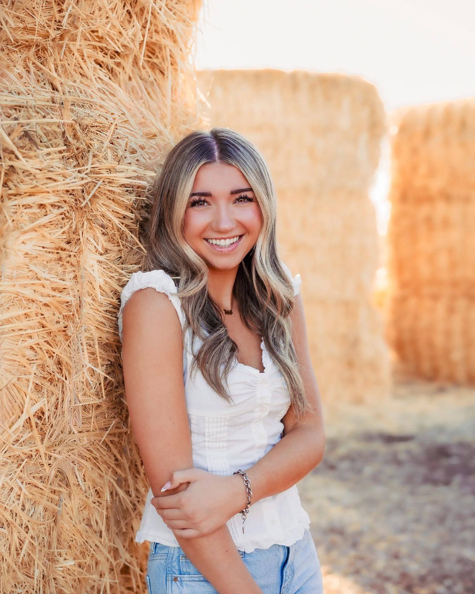 A smiling young woman with long blonde hair leaning against a stack of hay bales in a farm setting during sunset.