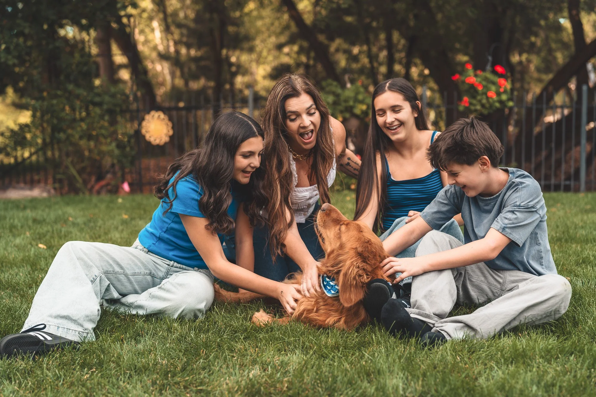 Four people and a dog sitting on grass in a backyard with trees and a fence. The group looks happy and engaged with the dog, which is a golden retriever puppy. It appears to be a warm, pleasant day.