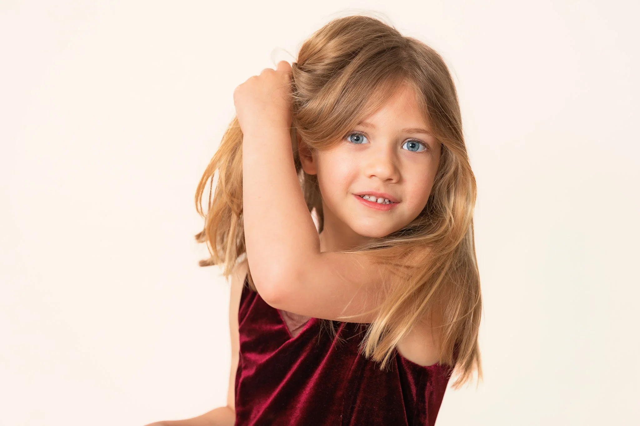 A young girl with long blonde hair and blue eyes, wearing a burgundy velvet top, smiling softly at the camera.