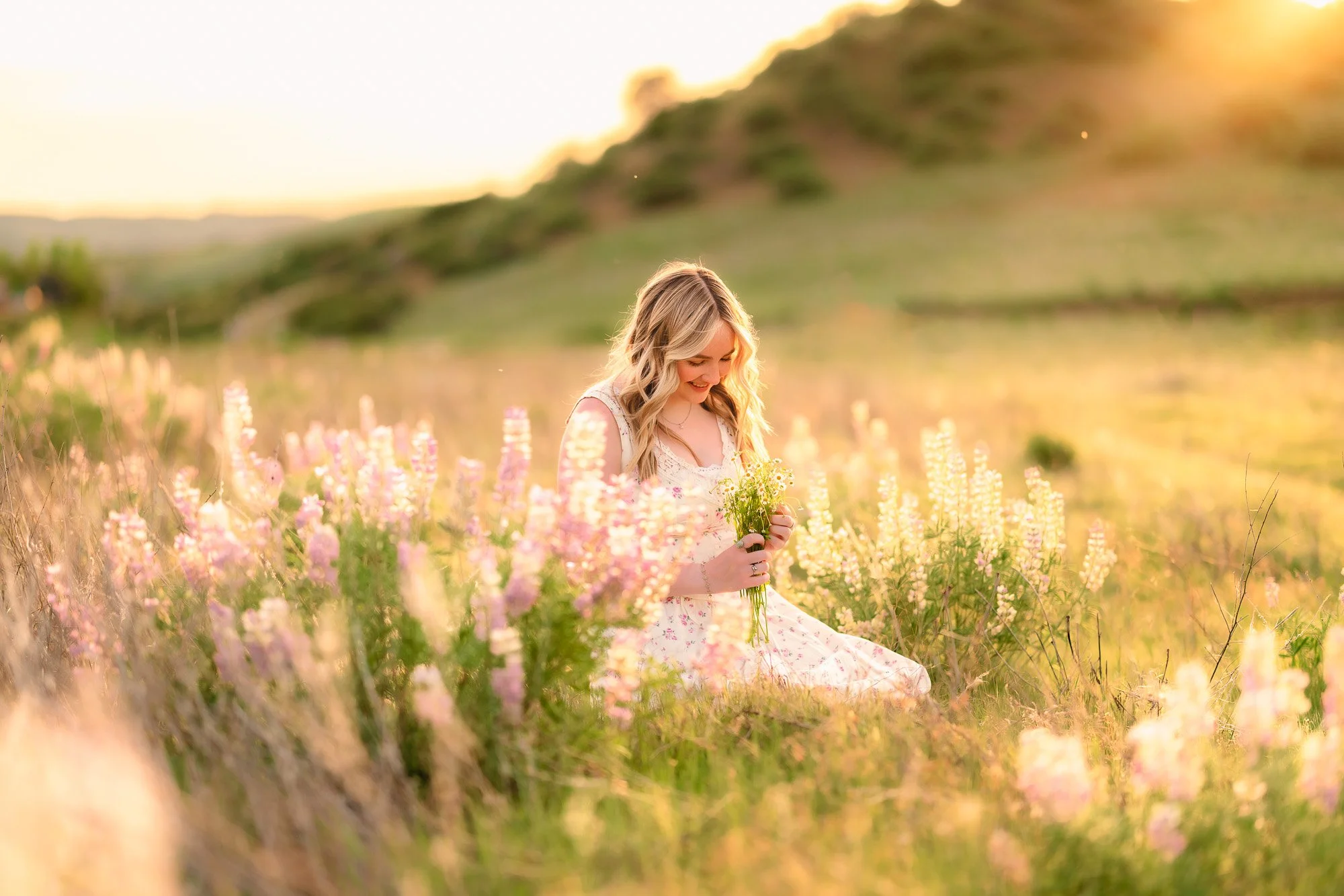 Young woman with blonde hair sitting in a field of pink and purple flowers during sunset, smiling and holding a small bouquet.