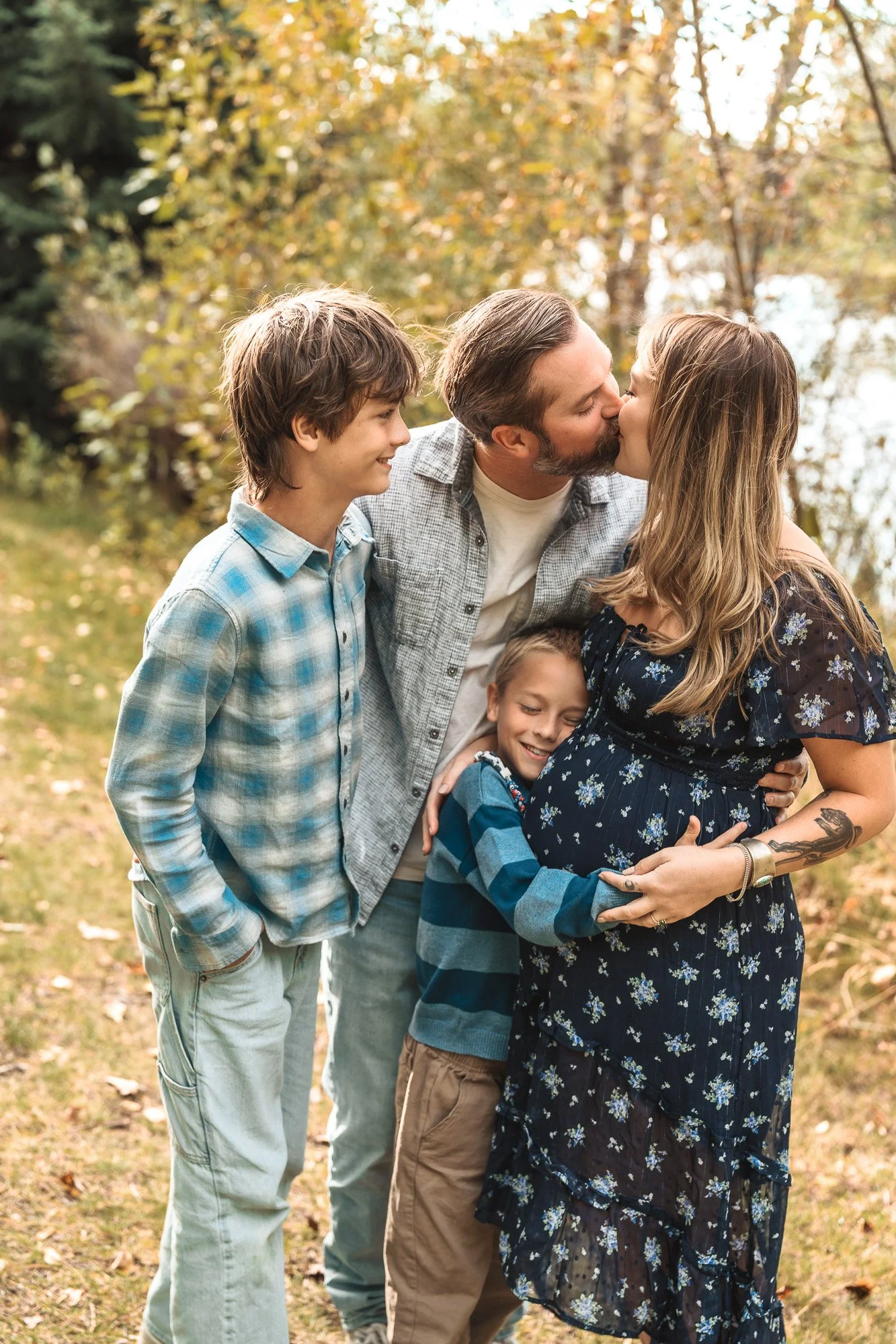 A family outdoors in a park during autumn, with trees and fallen leaves, as an adult man and woman kiss while two children, a boy and a girl, smile and hug each other.