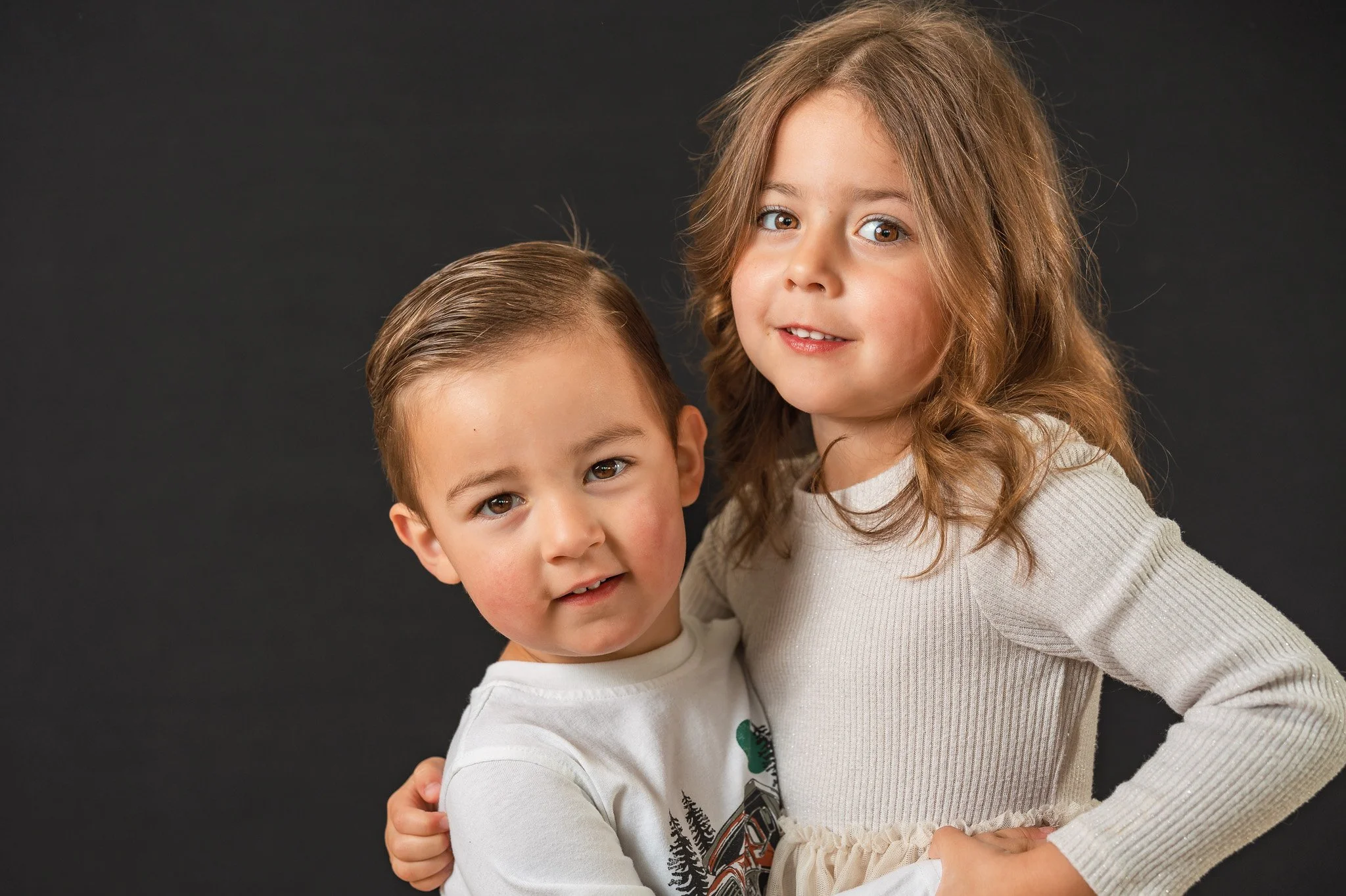 A young boy and girl standing close together against a black background, looking at the camera. The boy is hugging the girl, and they both have light skin and brown hair.
