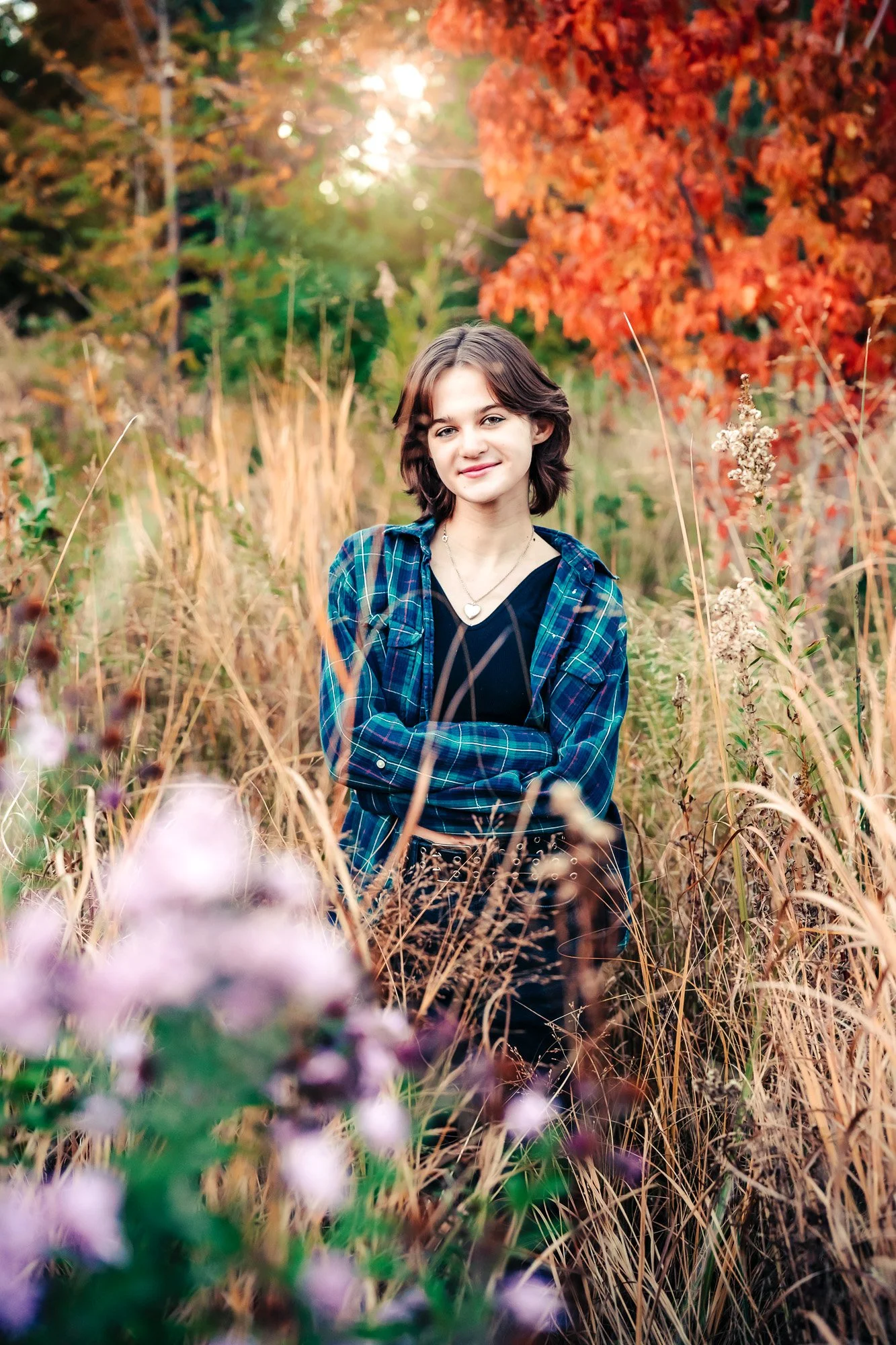 Young woman standing in a field of tall grass and wildflowers with autumn-colored trees in the background, smiling at the camera.