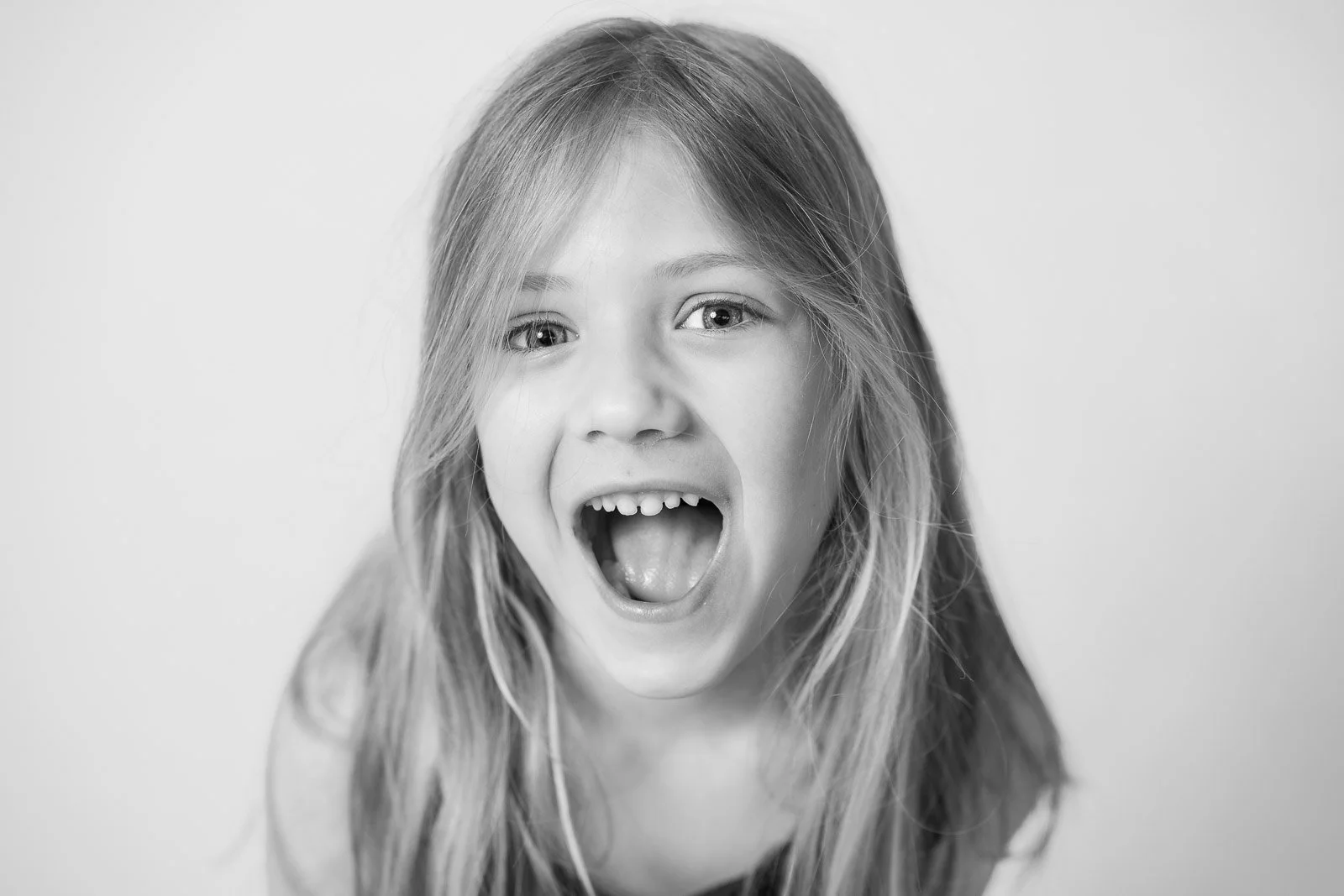 A young girl with long hair smiling with her mouth open, showing her teeth, in a black and white portrait.