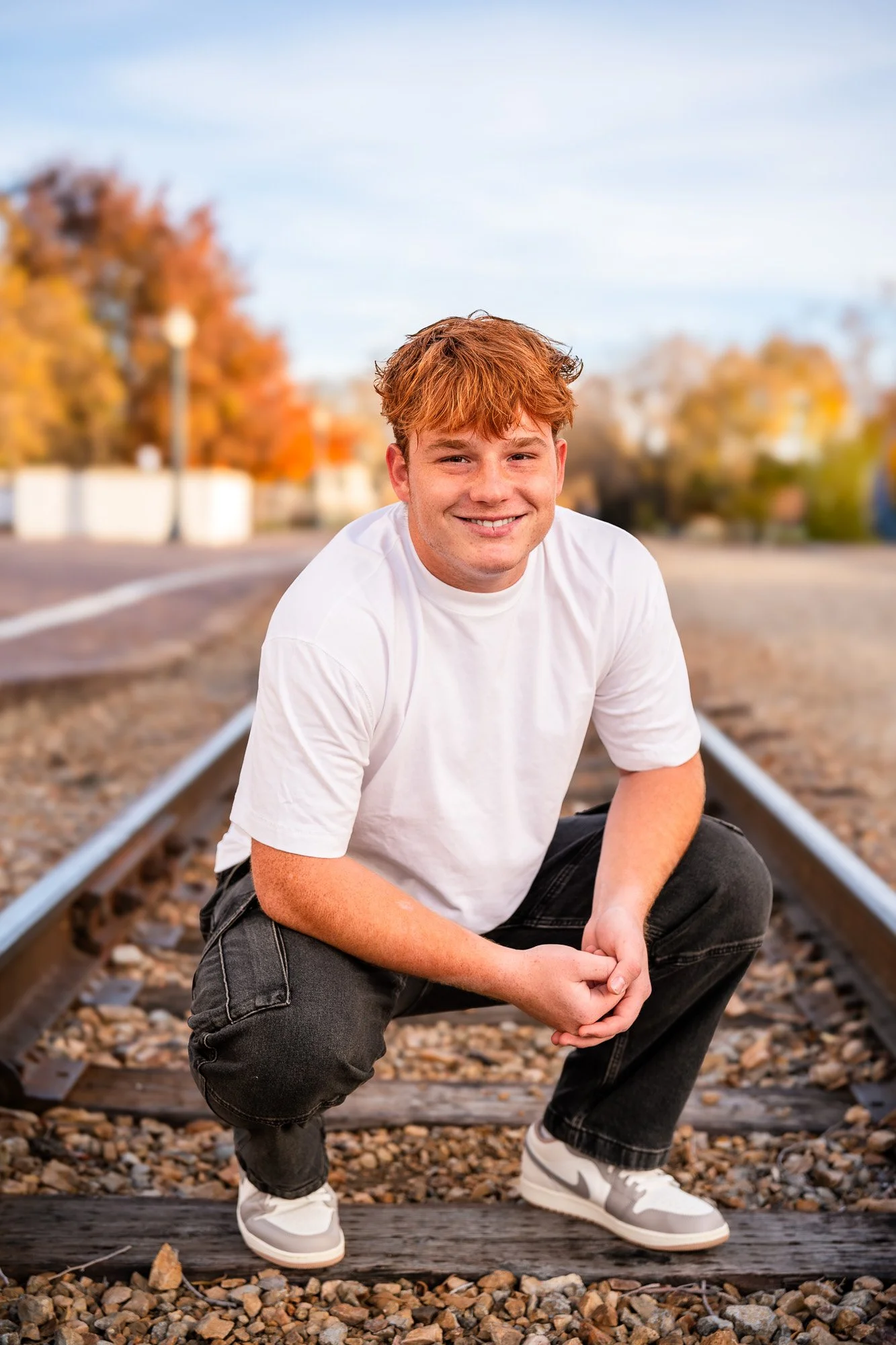 Young man with red hair and light skin crouching on train tracks, smiling, wearing a white t-shirt, black jeans, and white sneakers, with autumn trees in the background.