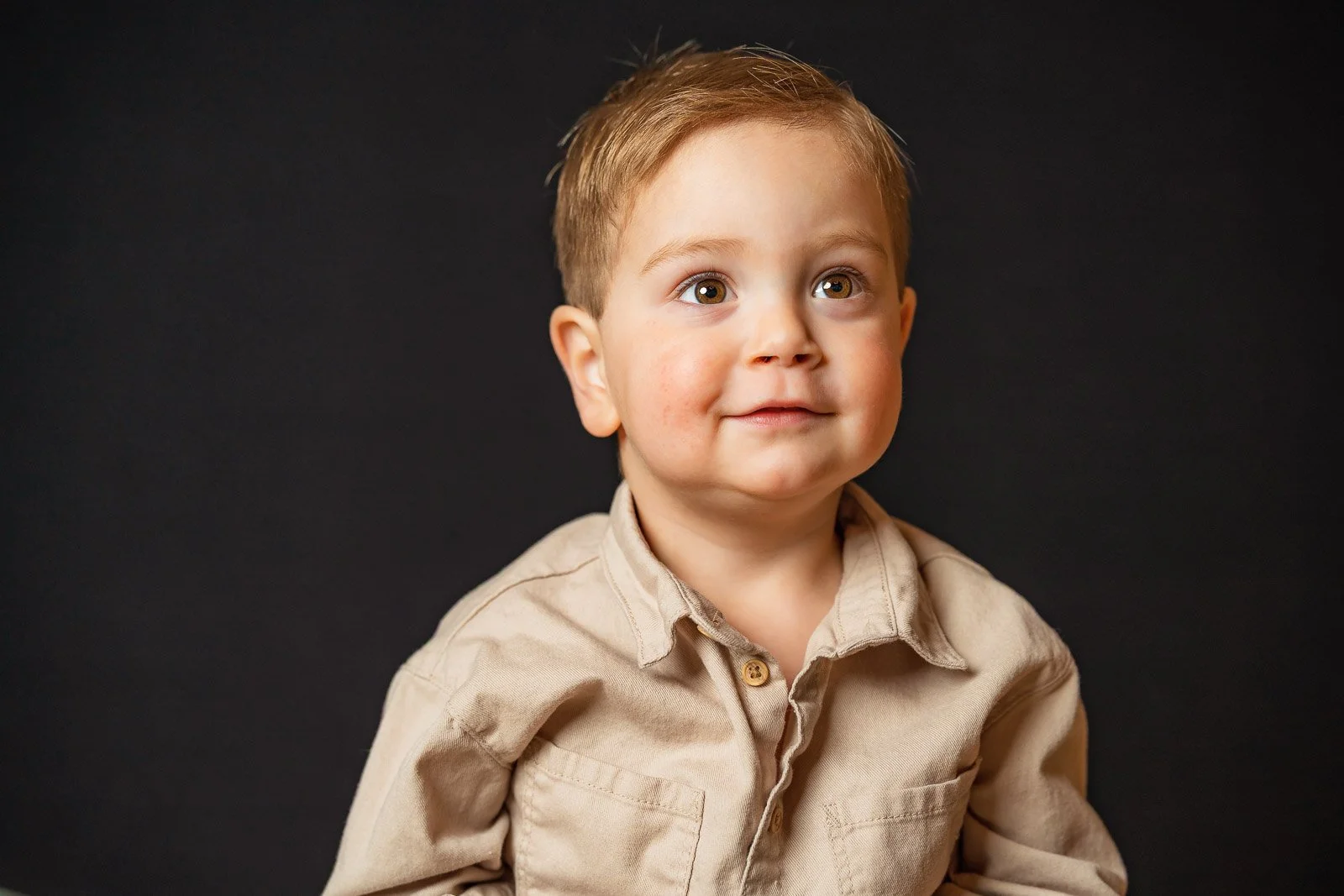 A young boy with light brown hair, brown eyes, and light skin, wearing a beige button-up shirt, looking slightly to his right with a neutral expression, against a dark background.