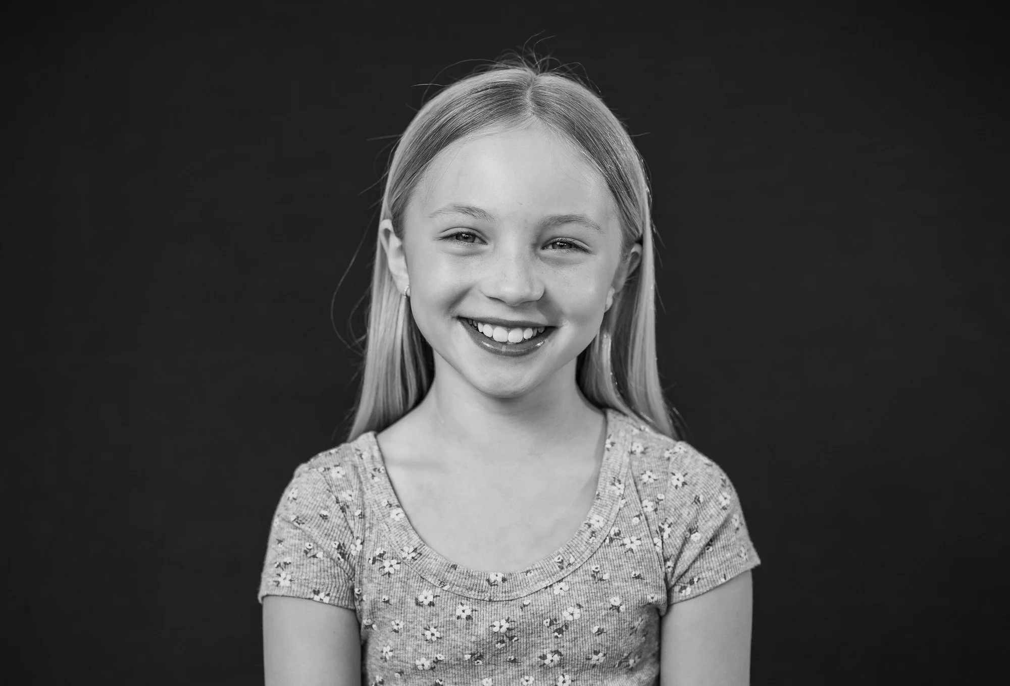A young girl smiling, with long hair, wearing a floral patterned shirt, black and white photo, against a dark background.