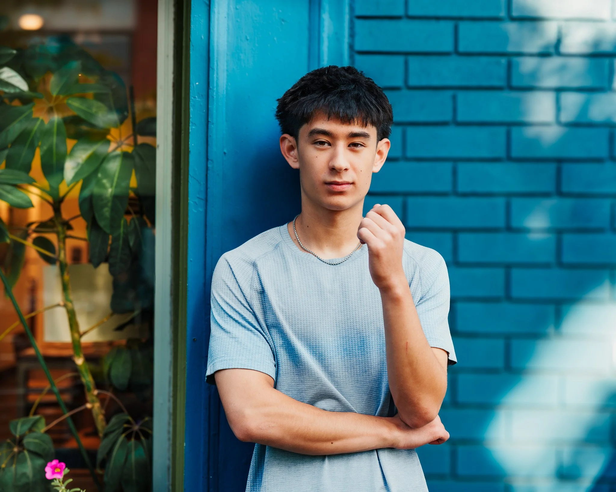 Teenage boy with short black hair in light gray t-shirt and silver chain leaning against blue brick wall during downtown Boise male senior pictures session by Eagle senior photographer.
