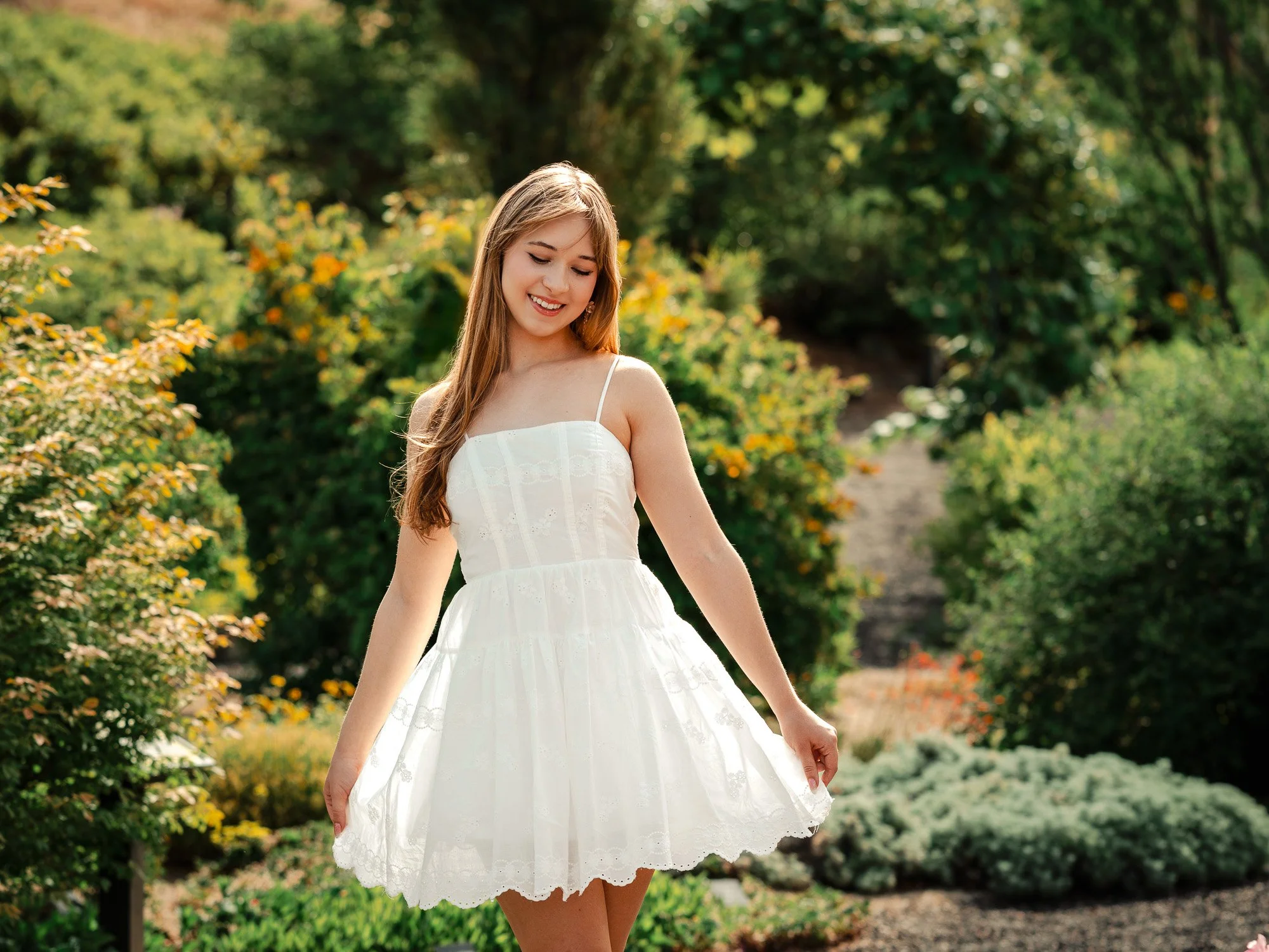 A young woman in a white summer dress standing outdoors among greenery and flowers, smiling and looking down.