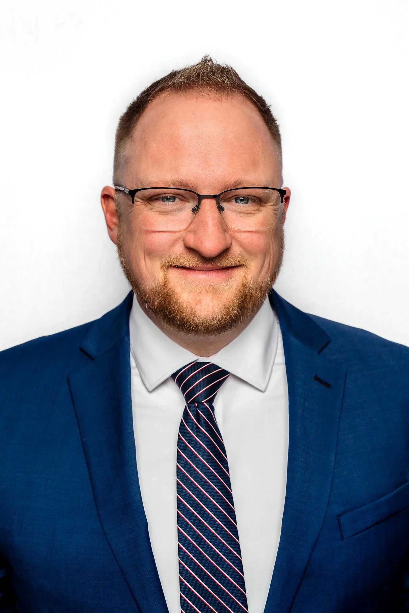 A man with glasses and a beard wearing a blue suit, white shirt, and striped tie, smiling against a plain white background.