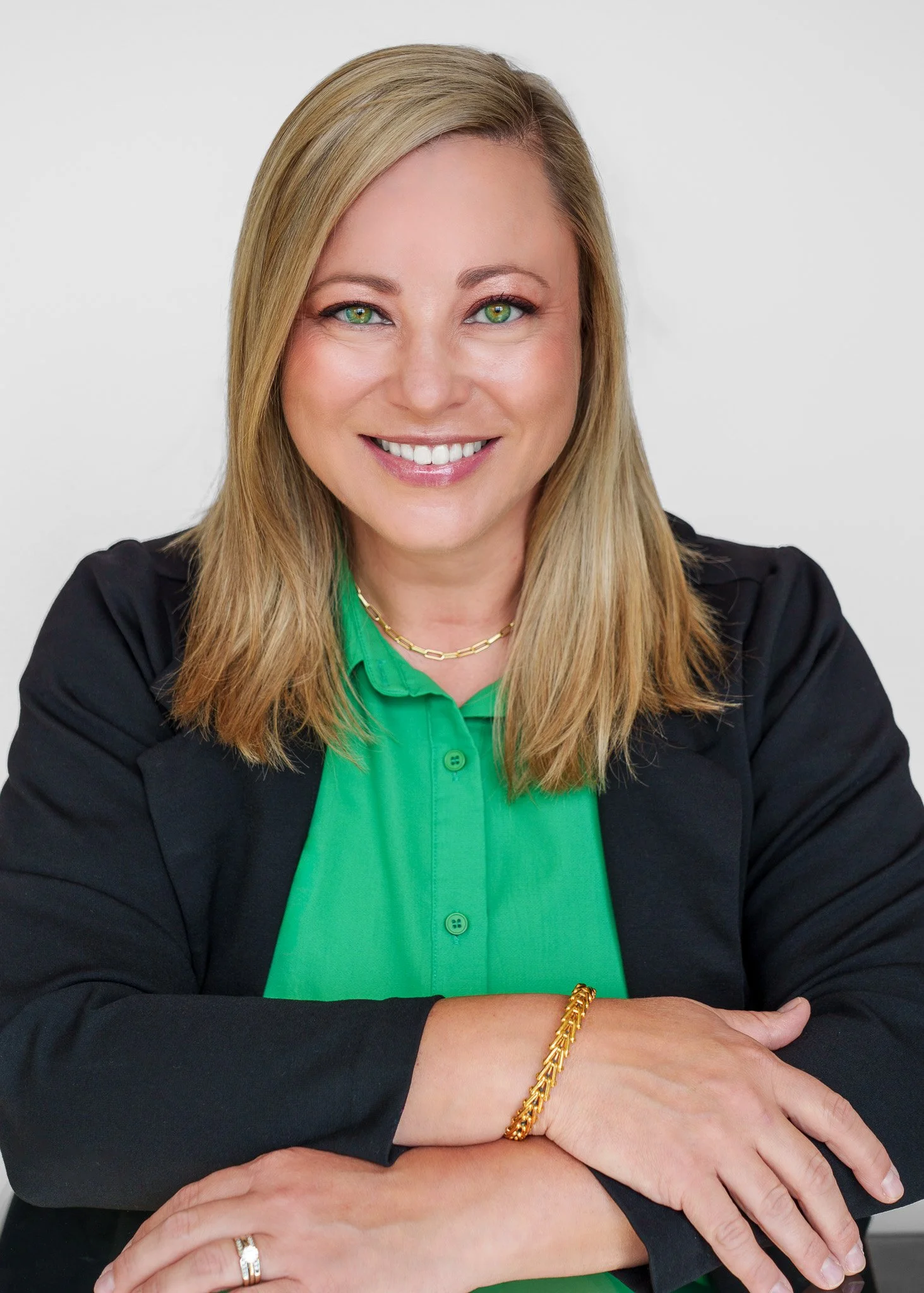 Portrait of a smiling woman with blonde hair, wearing a black blazer, green blouse, a gold chain necklace, and a gold bracelet, sitting with her arms crossed, against a plain light background.