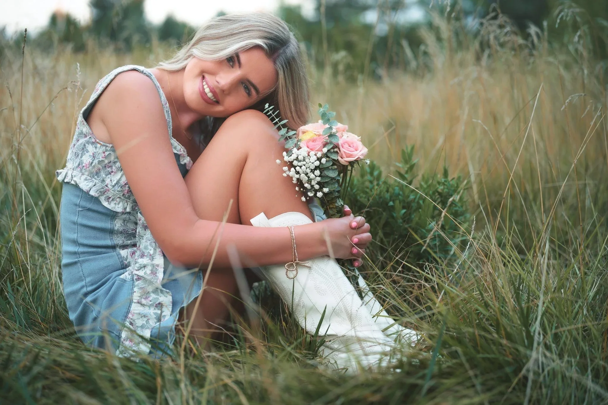 A woman sitting on the grass in a field, smiling while holding a bouquet of pink roses and white baby's breath flowers.
