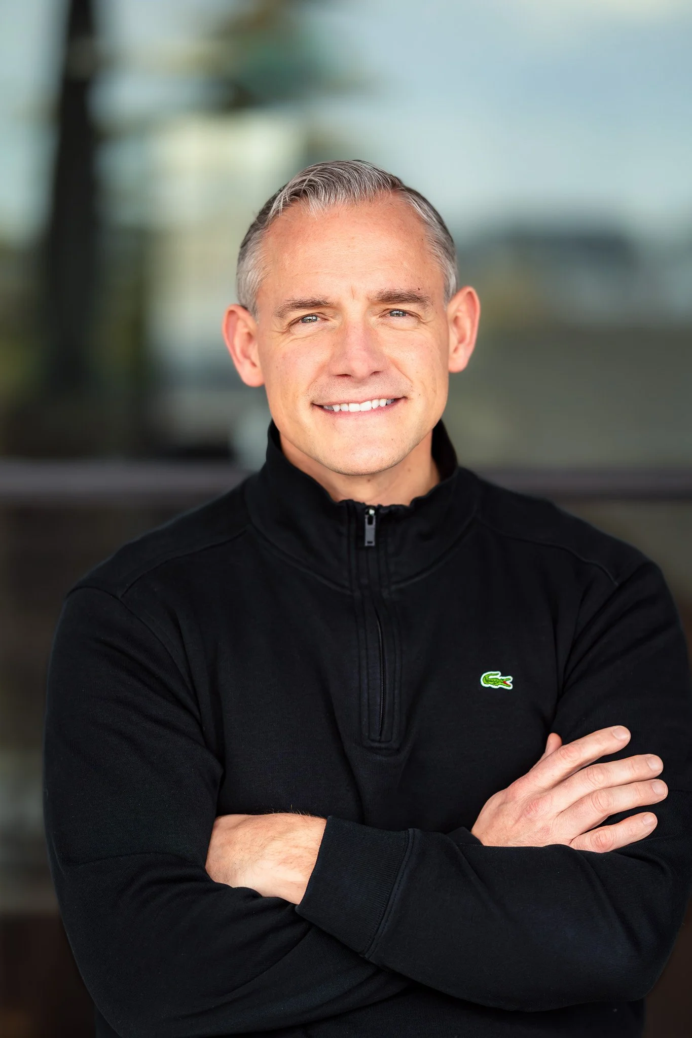 A man with short gray hair, wearing a black jacket with a green crocodile logo, standing with arms crossed and smiling outdoors.