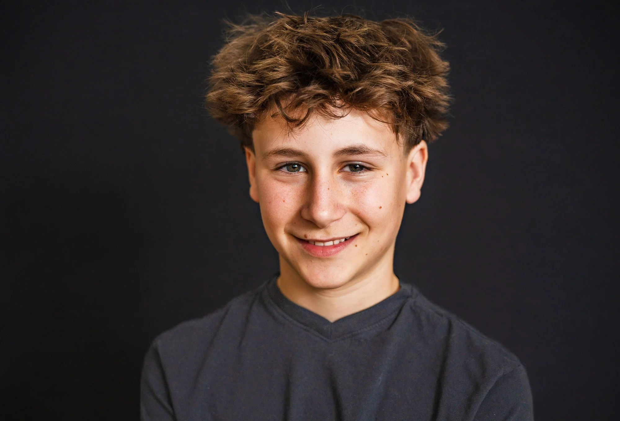 A teenage boy with curly brown hair, blue eyes, and a light complexion smiling against a dark background.