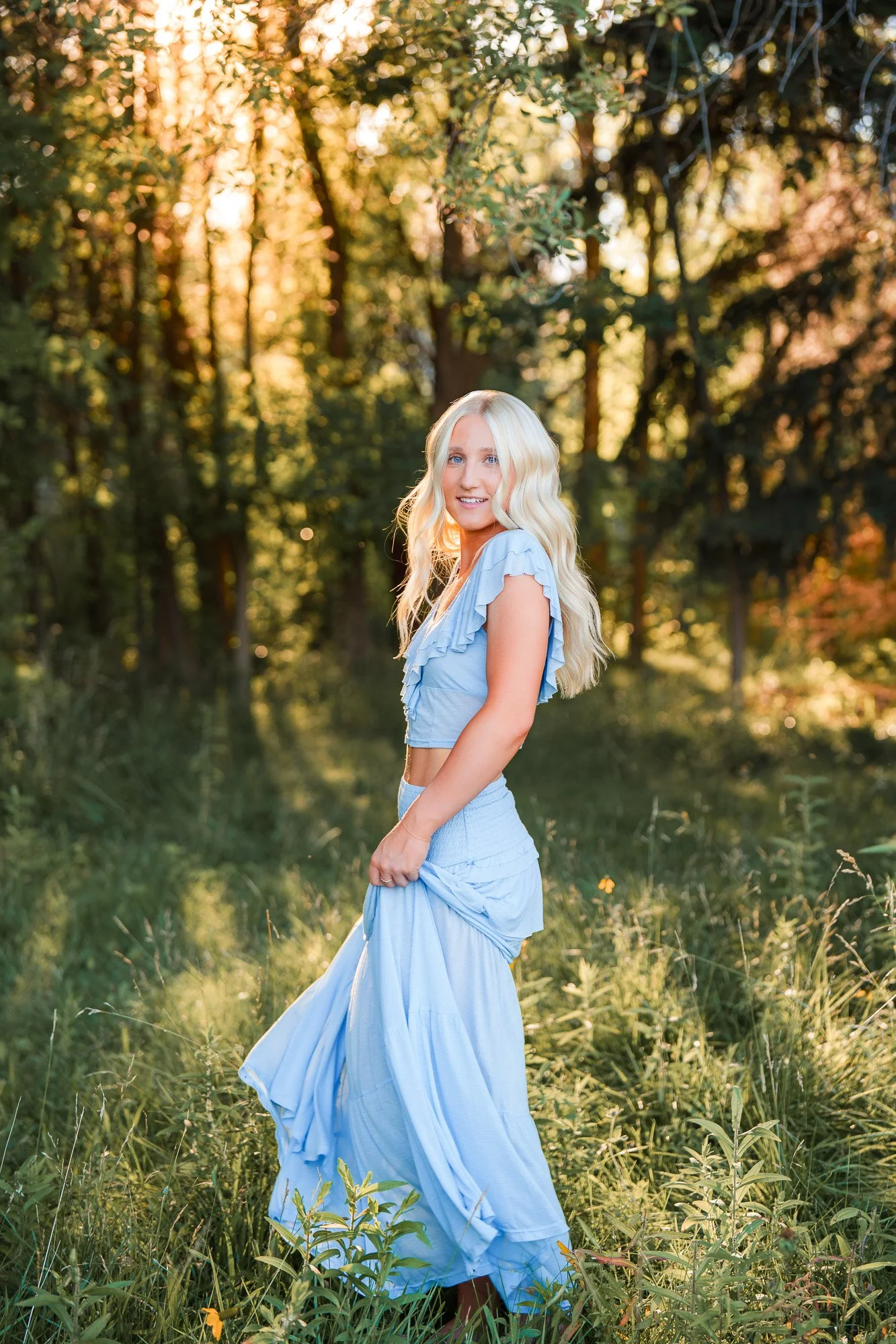 A young woman with long blonde hair wearing a blue dress, smiling and standing in a grassy, wooded area during sunset.