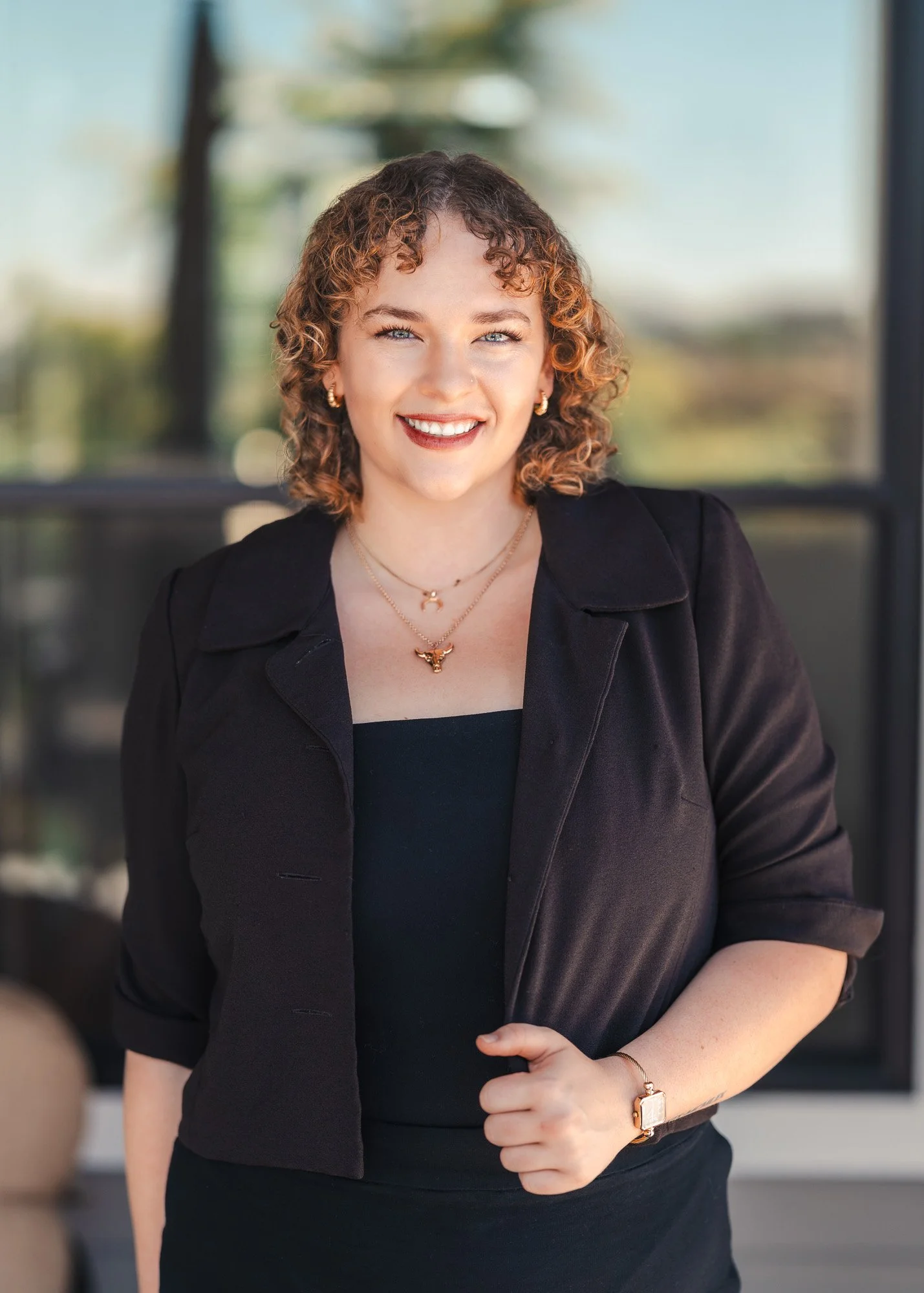 A woman with short curly hair and blue eyes, smiling, wearing a black blazer and a black top, accessorized with gold jewelry and a rose gold watch, standing outdoors with a blurred background.