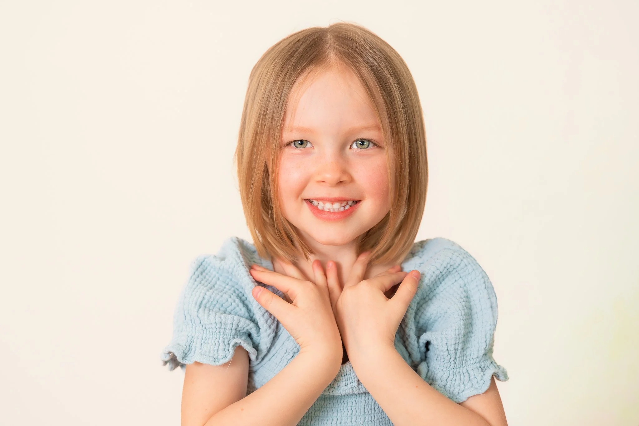 A young girl with shoulder-length red hair, blue eyes, and freckles, wearing a light blue ruffled shirt, smiling and touching her neck with both hands.