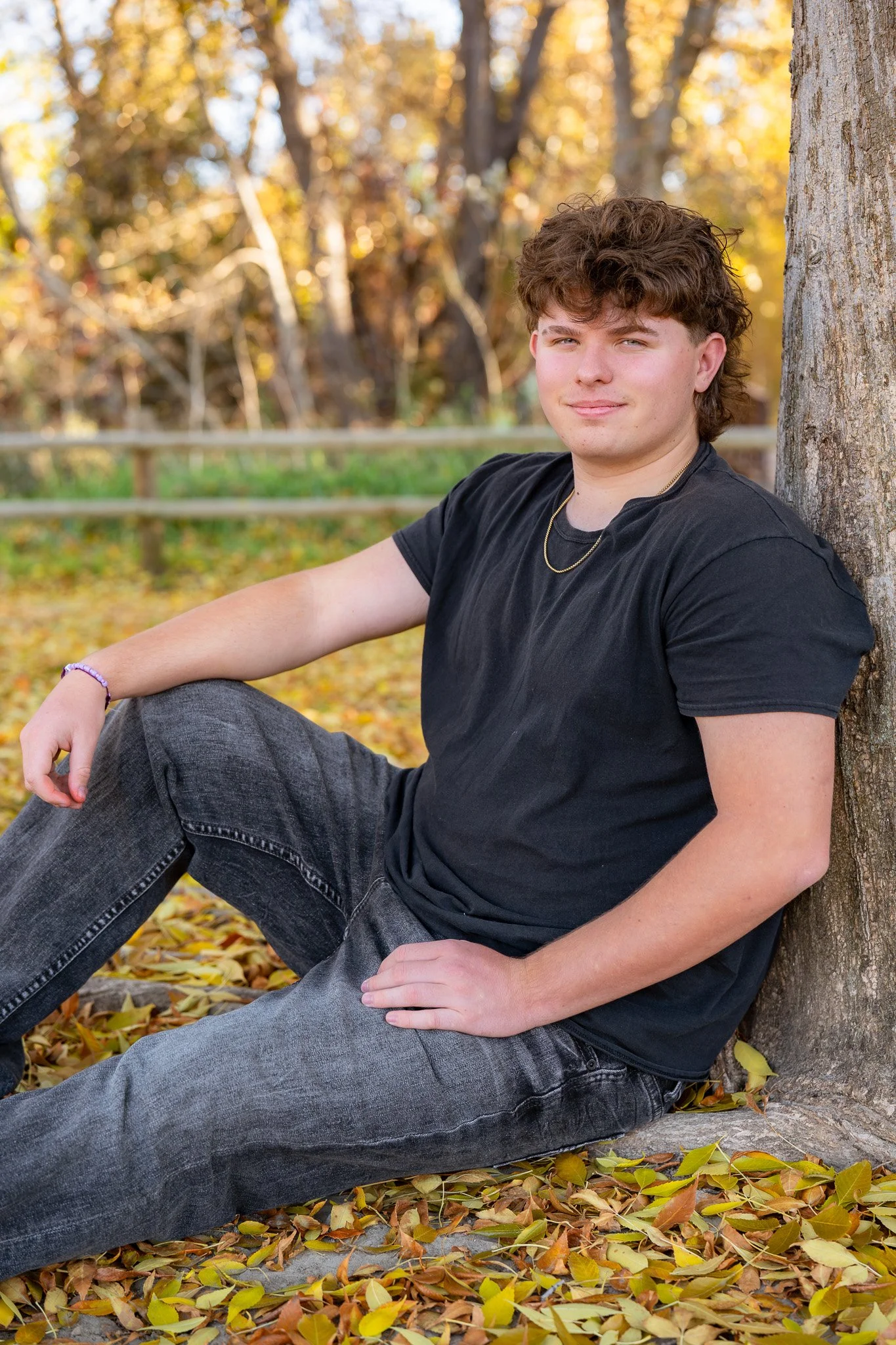 Young man sitting against a tree during his senior photo session in Eagle, Idaho fall, surrounded by colorful fallen autumn leaves.