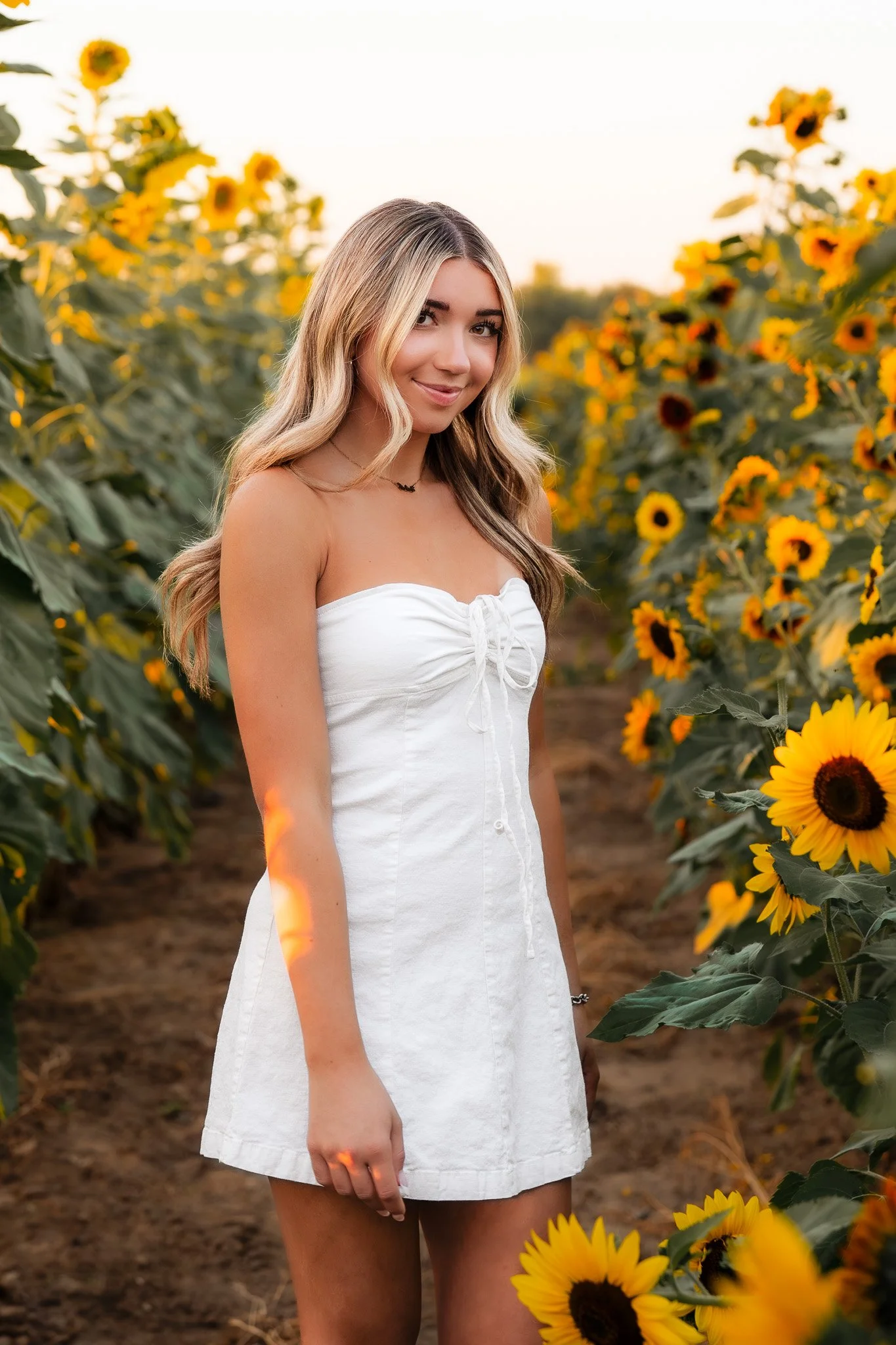 A young woman with long wavy blonde hair stands in a sunflower field wearing a white strapless dress and smiling.