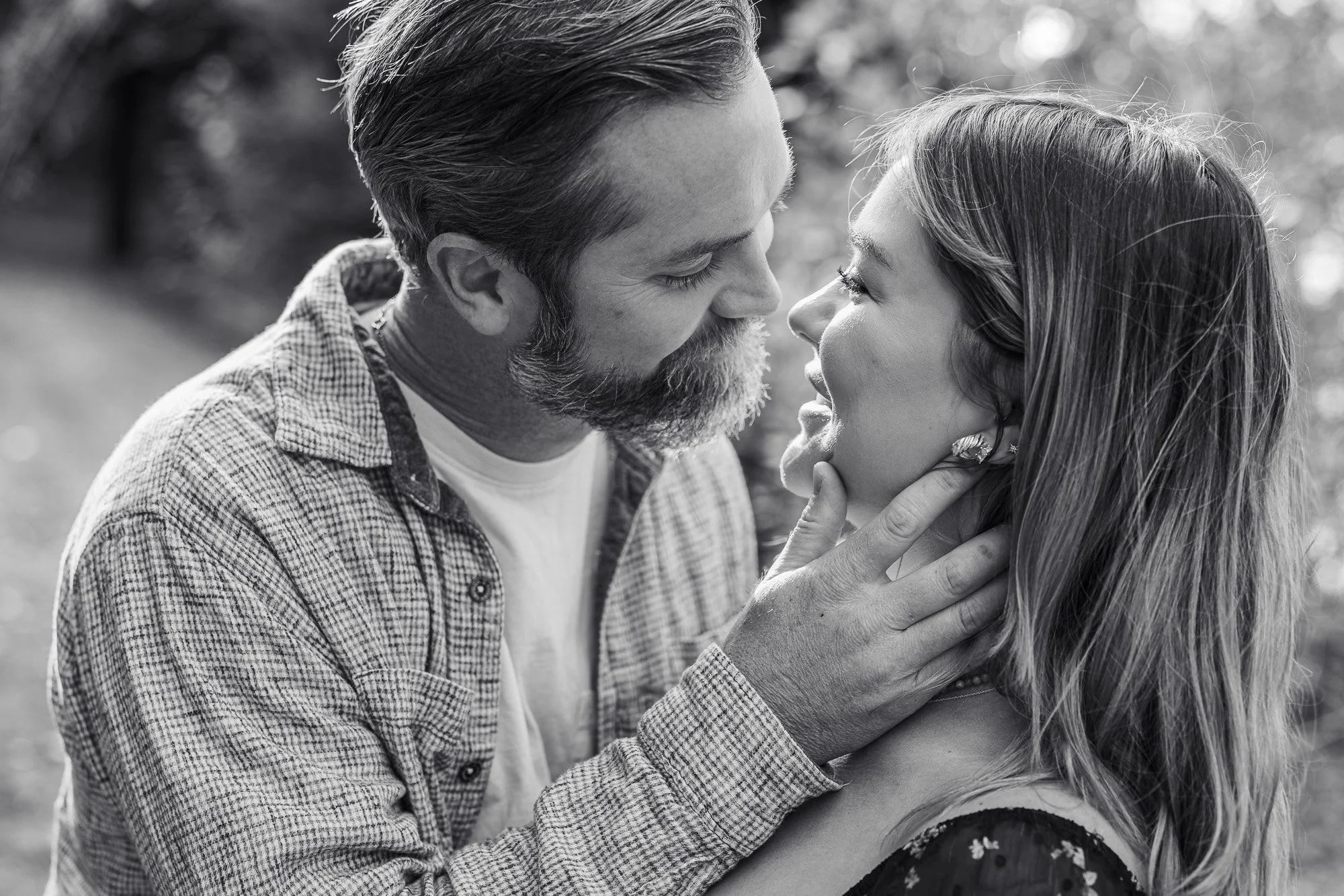 A black and white photo of a man and woman close together, touching faces and gazing into each other's eyes outdoors.
