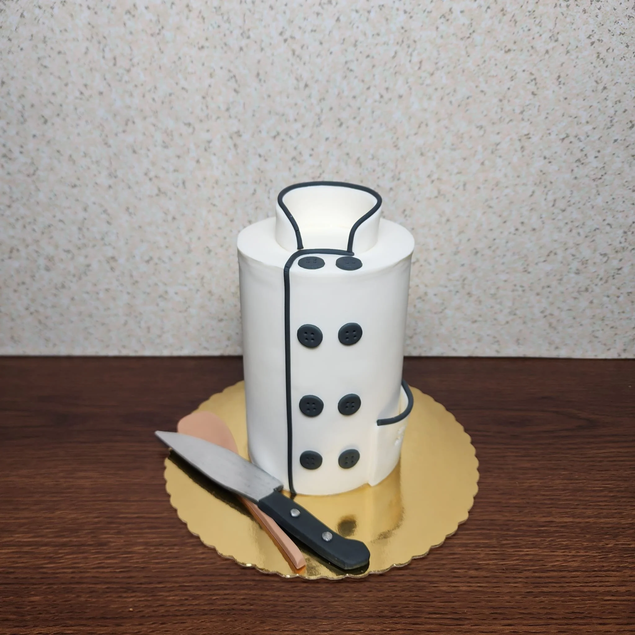 Pastel con diseño de chaqueta de chef y un cuchillo negro sobre una base dorada, en una mesa de madera, con fondo de papel decorativo.