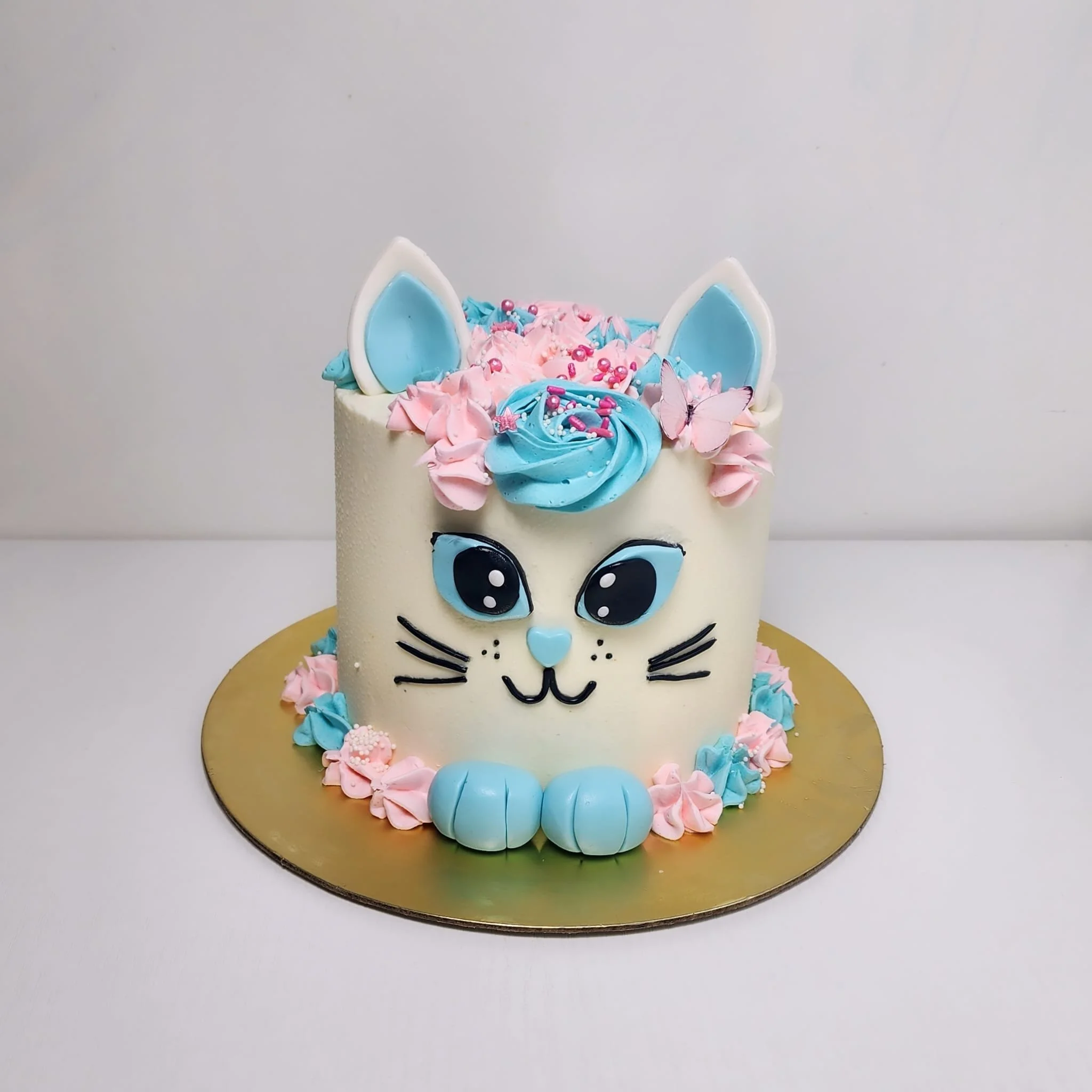 Pastel de cumpleaños con temática de gato de color blanco, orejas azules, grandes ojos azules, cara dibujada con glaseado y decoración en rosa y azul con mariposas y confites