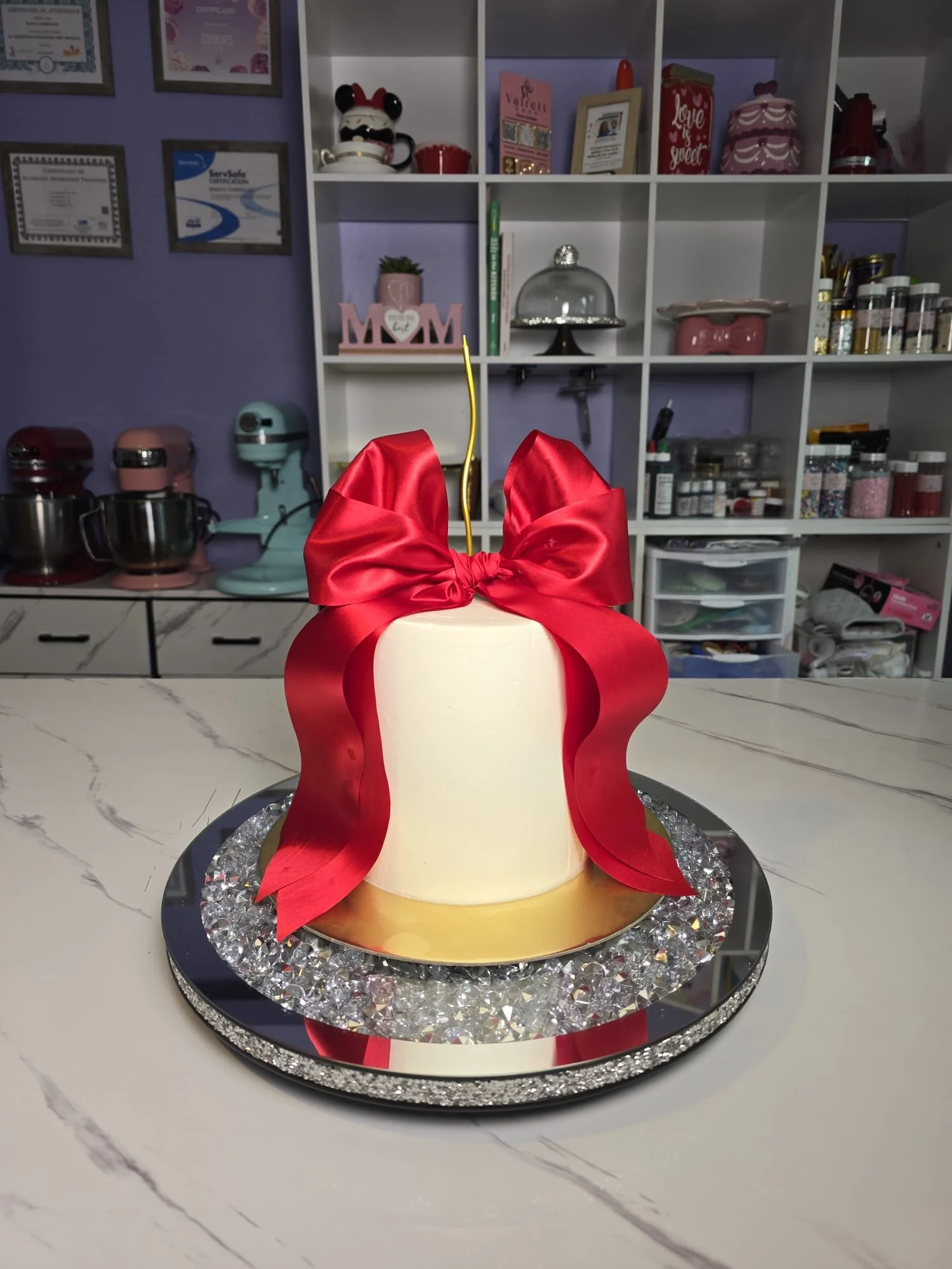 Pastel de cumpleaños blanco decorado con una gran lazo rojo en la parte superior, colocado en una bandeja con apariencia de diamantes y espejo, sobre una mesa de mármol.