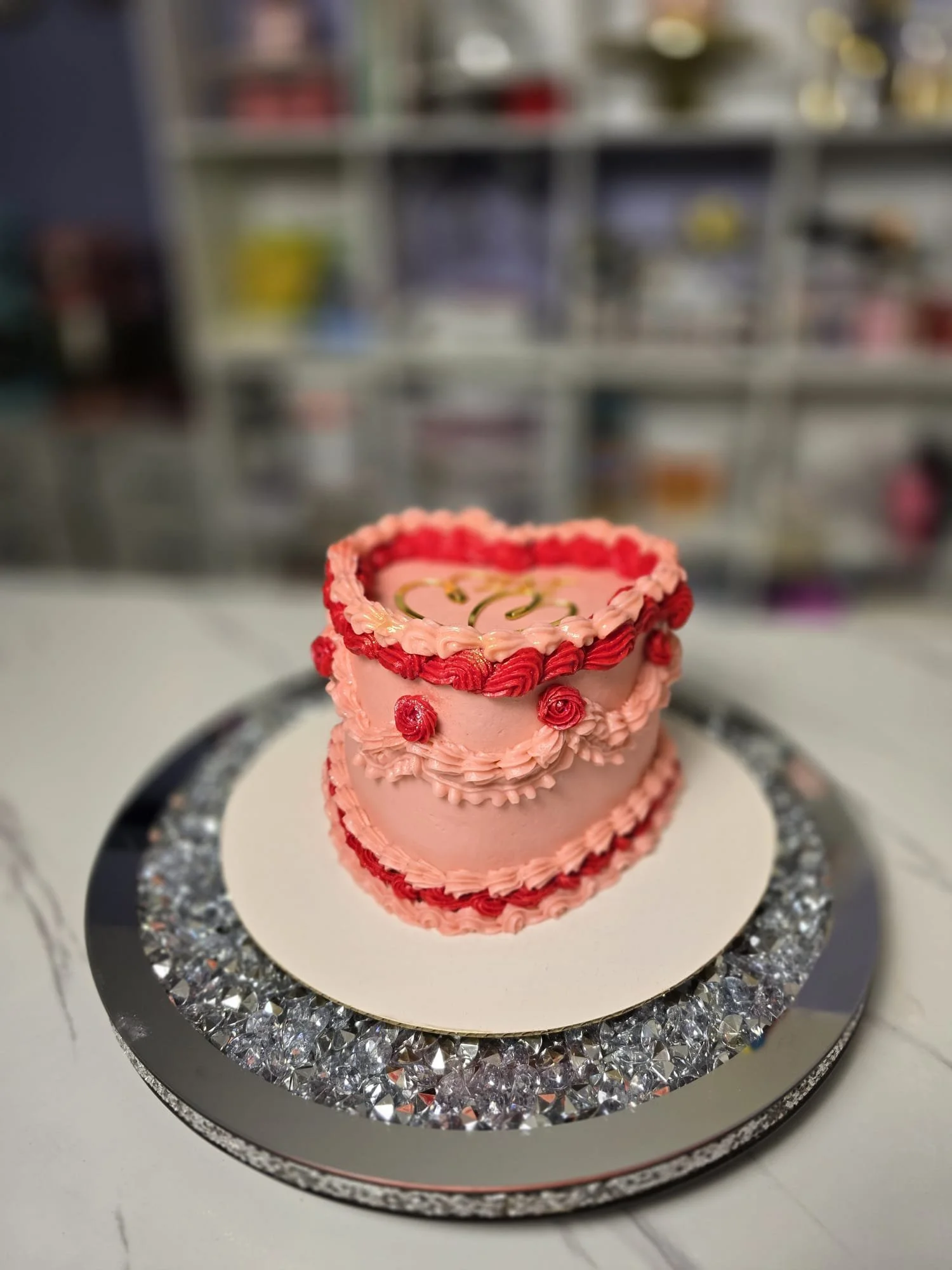 Pastel de dos pisos decorado con glaseado rosa y rojo, con rosas de decoración, sobre un plato de espejito y fondo de estantería con artículos variados.