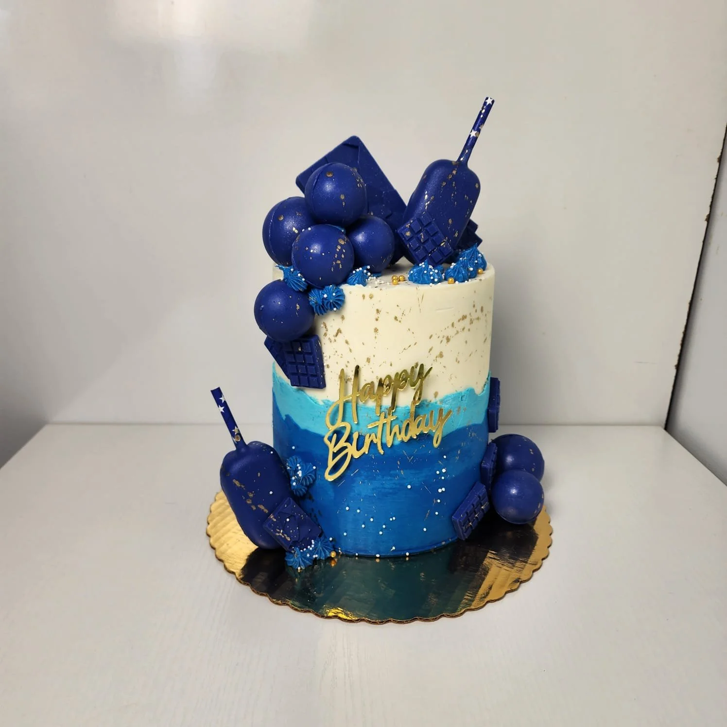 Pastel de cumpleaños decorado en tonos azules y blancos con detalles en dorado, con bolas, chocolates y una leyenda que dice 'Happy Birthday'.