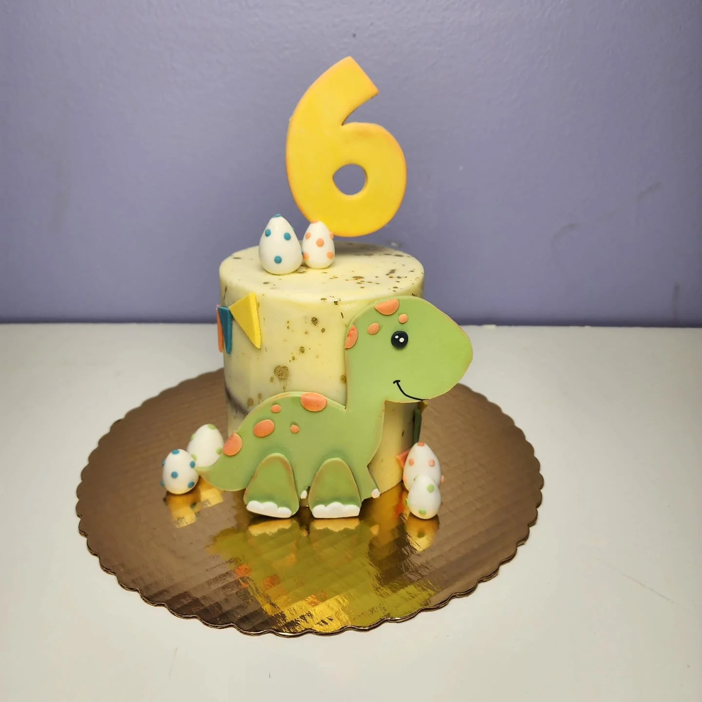 Pastel de cumpleaños con figura de dinosaurio y decoraciones, incluyendo un número 6 amarillo.