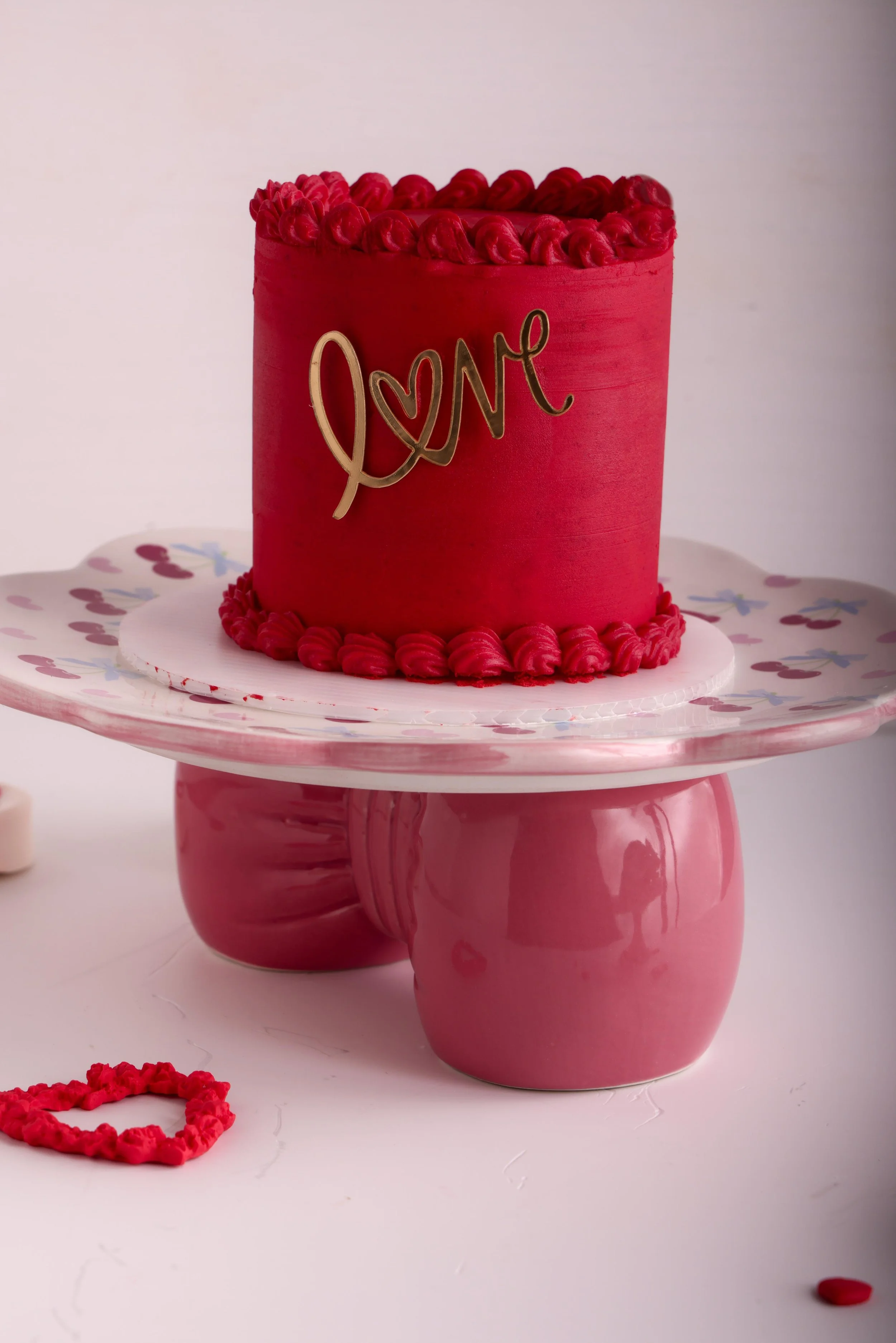 Pastel de cumpleaños rojo con la palabra 'Love' en letras doradas, decorado con crema en la parte superior, sobre un soporte rosa con patrones y rodeado de pequeños corazones de azúcar.