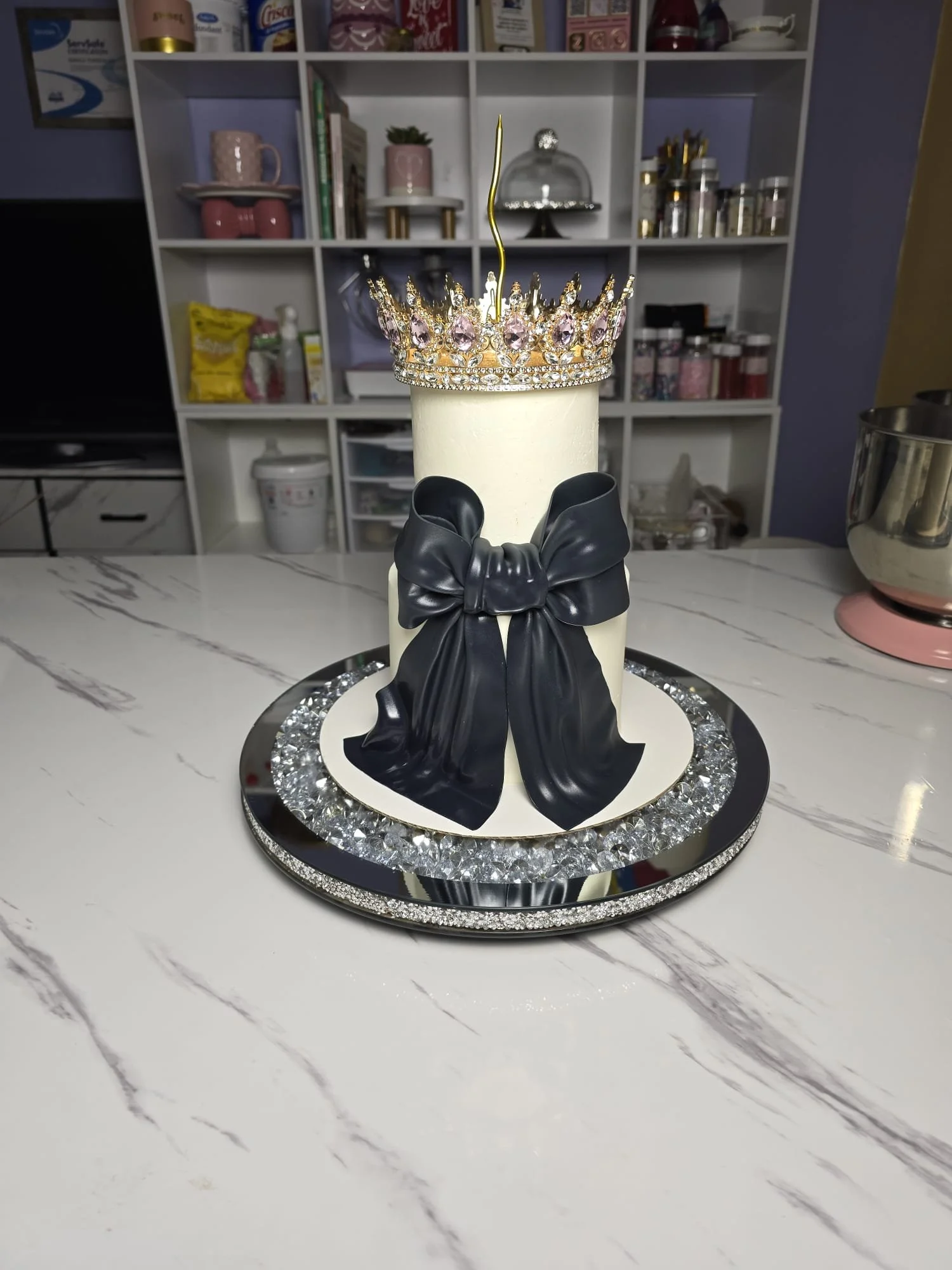Pastel decorado con una corona, un lazo negro y una vela blanca en forma de taza, sobre una base brillante con cristales.